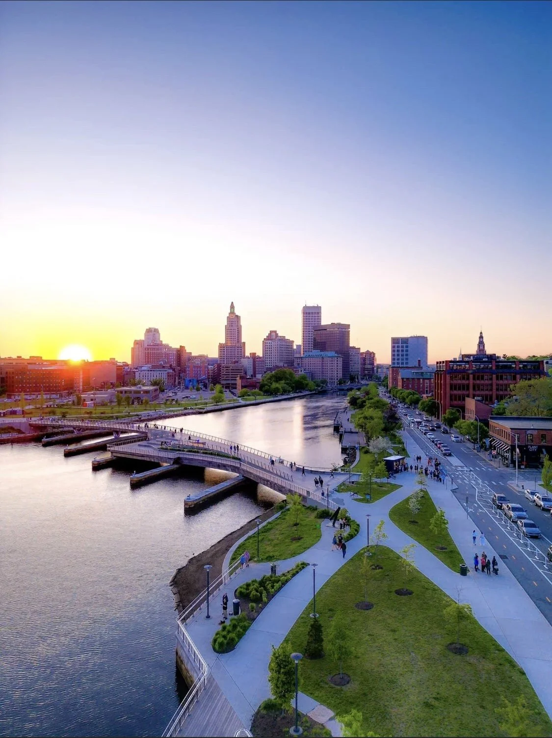 City skyline at sunset with a river and a park in the foreground.