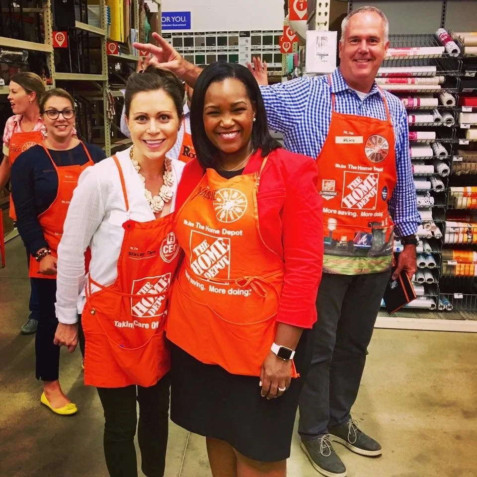 A group of five smiling people at a Home Depot store, all wearing orange aprons, likely participating in a store event or team activity. Stacey Tank, Yanique Woodall, Mike Hibbison.