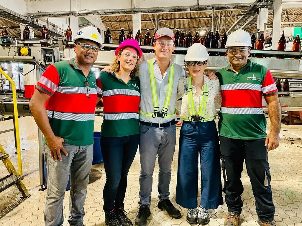 Five HEINEKEN colleagues including Stacey Tank in safety gear posing together in an industrial setting, with brown bottles on a conveyor belt in the background in Surinam.