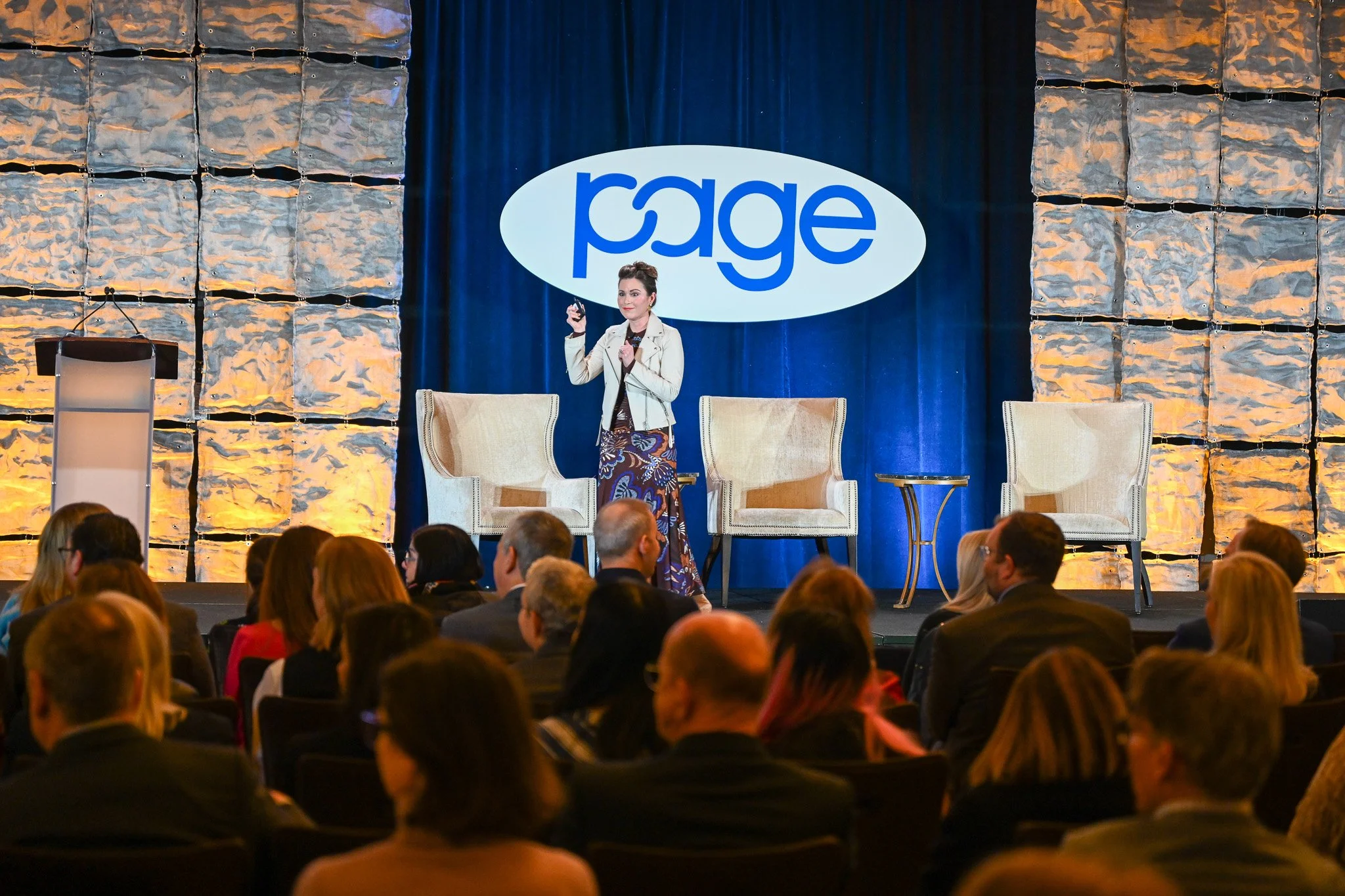 A woman is standing on stage at a conference, holding a device, with an audience watching. Behind her, there's a large banner with the word 'Page' in a blue logo, and two chairs are positioned on the stage.