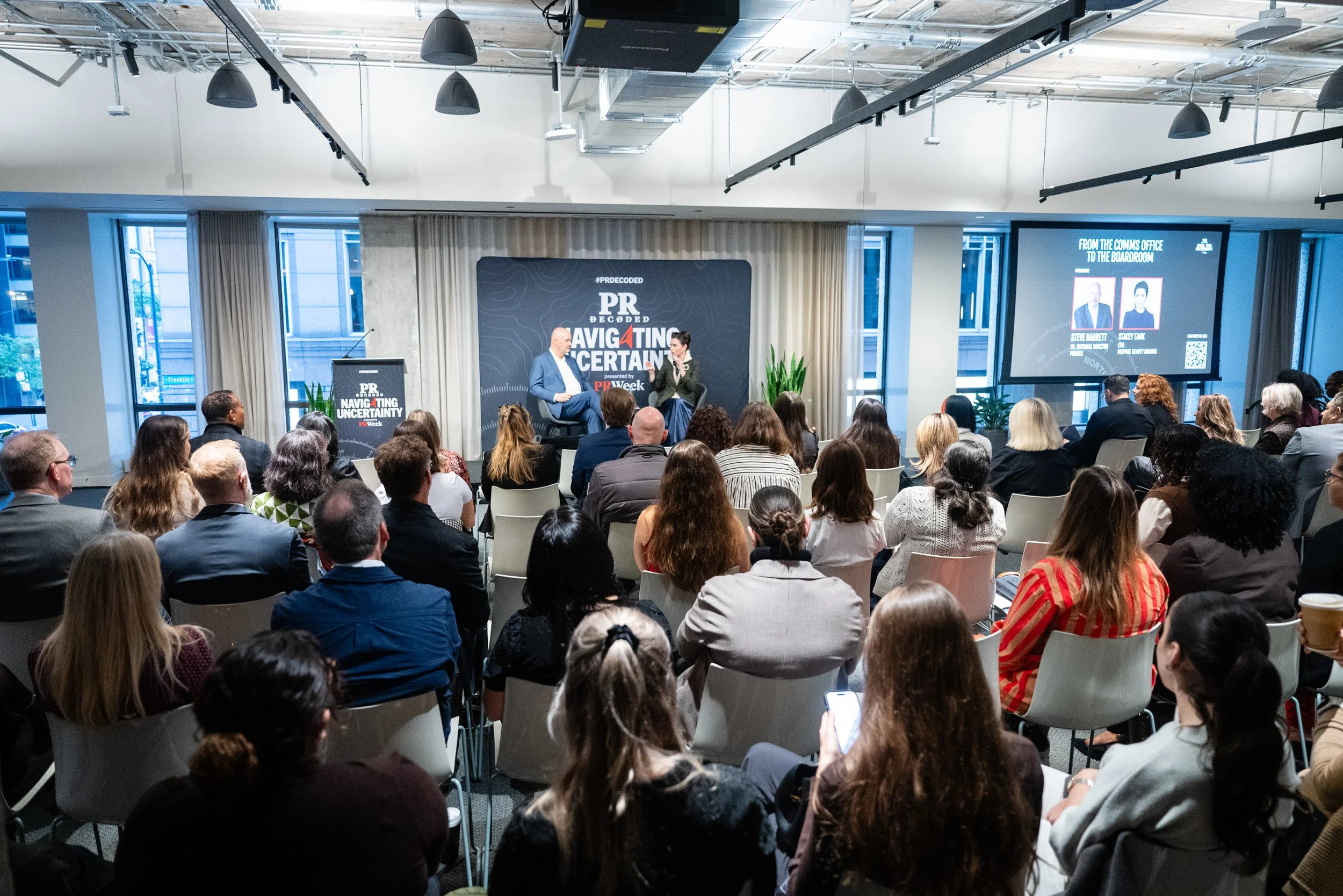 A large group of people attending a conference or seminar in a modern, well-lit room with large windows. A stage with two speakers, including Stacey Tank, and presentation screens displays the event's theme, 'PR Decoded: Navigating Uncertainty.'