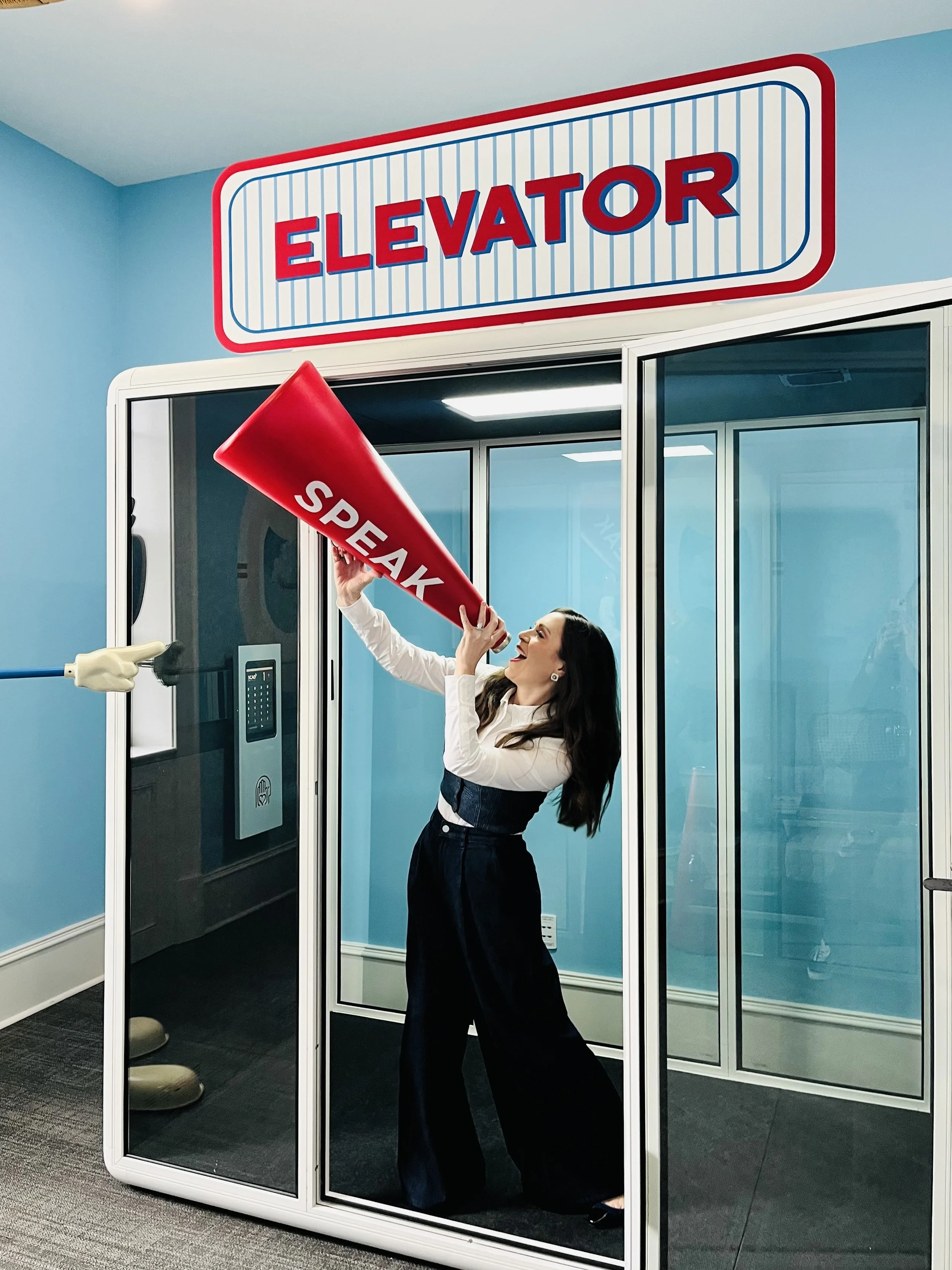 CEO and board director Stacey Tank standing inside a glass booth, holding a red megaphone labeled 'SPEAK' and smiling. The booth is marked 'ELEVATOR' on a sign above. Savannah College of Art & Design. SCAD.