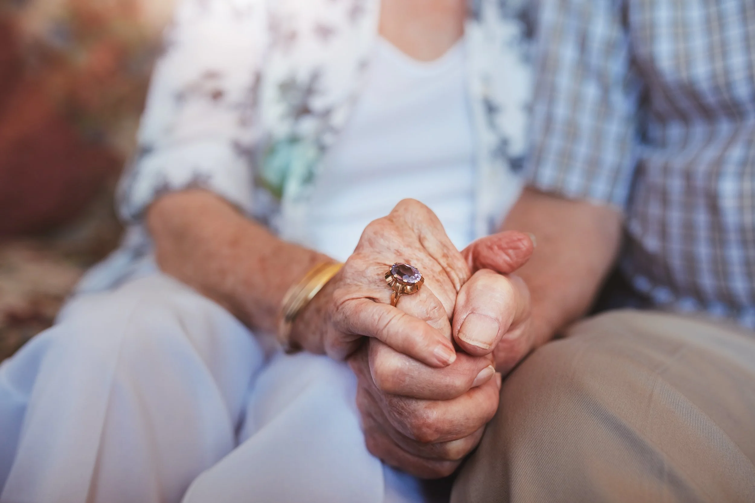 Close-up of an elderly person's hand with a large gemstone ring, being gently held by another person's hand, with a blurred background of people wearing casual clothing.
