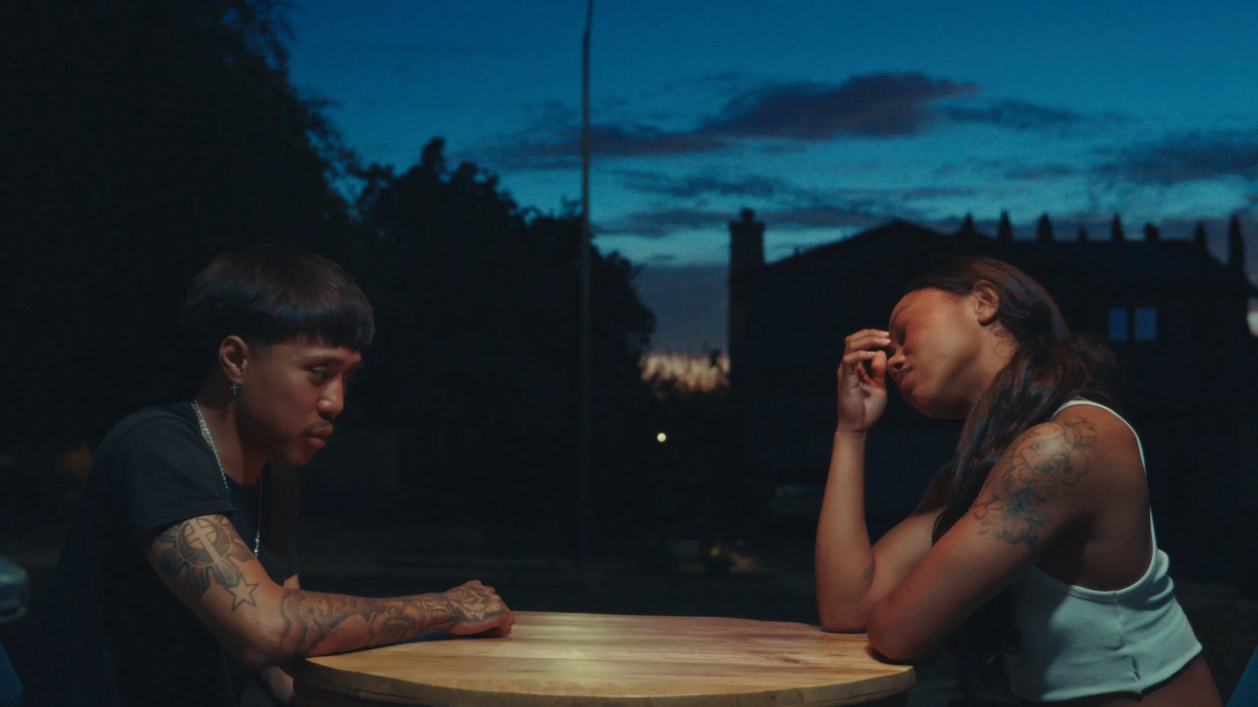 Two women with tattoos, sitting across from each other at a table outdoors during dusk, engaged in a moment of emotional vulnerability.