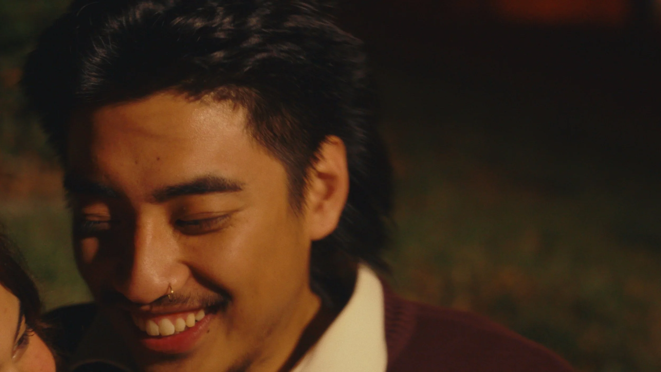 Close-up of a smiling young man with dark hair and facial hair, looking downward with his eyes closed.