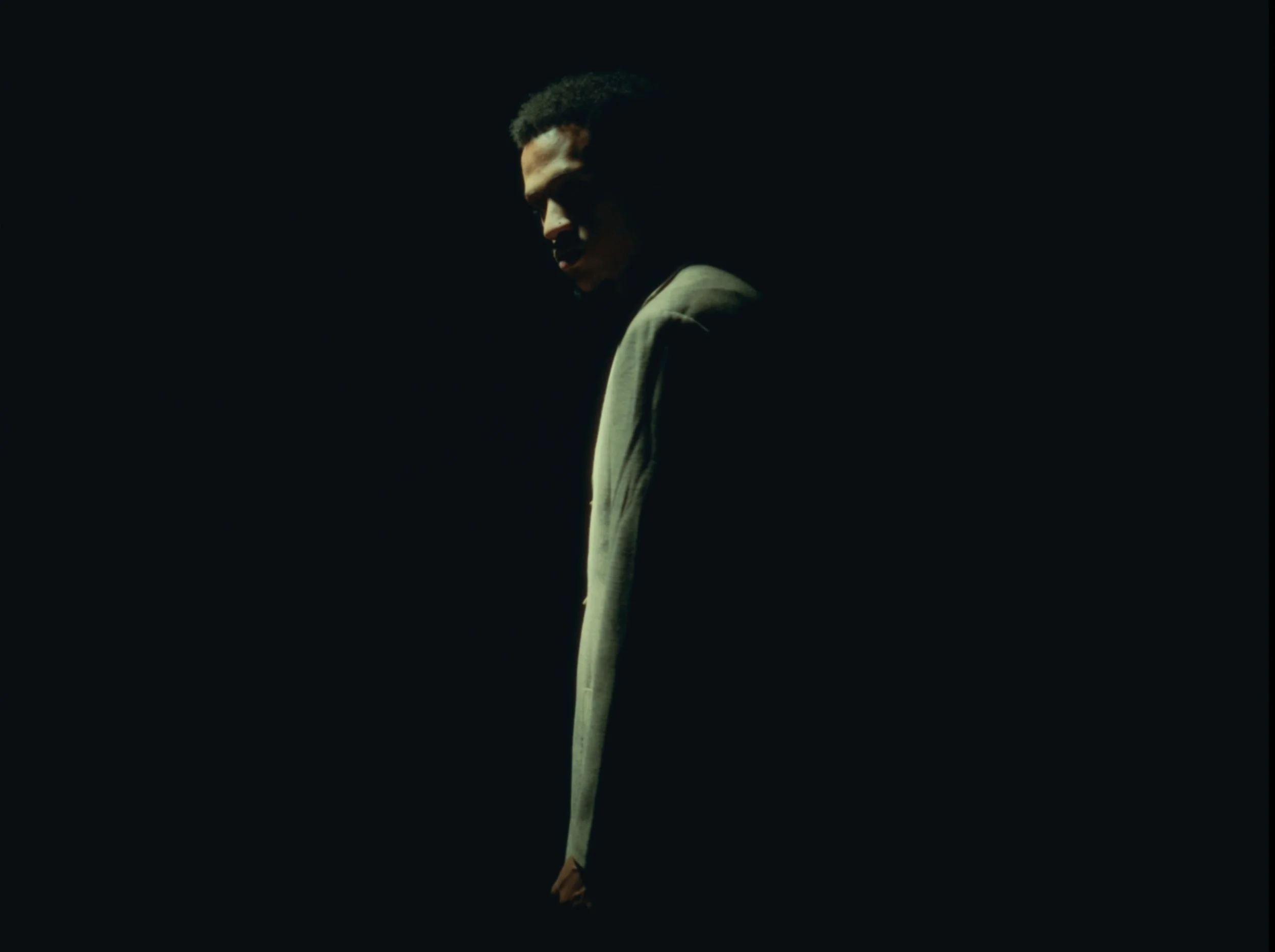 A dark background with a partial profile of a person with curly hair, facing downward, illuminated by a soft light, with part of their face and a light-colored shirt visible.
