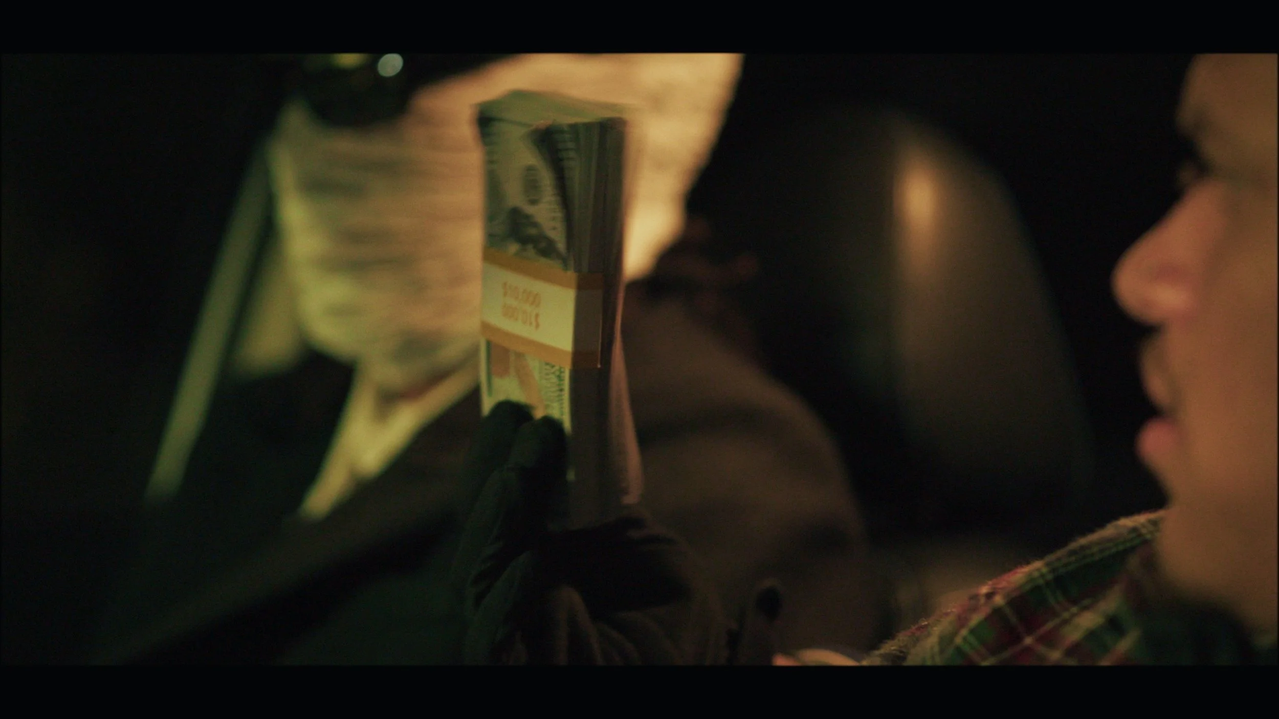A person holding a stack of money and a credit card, with a person's face visible on the right side of the image. The scene is dimly lit.