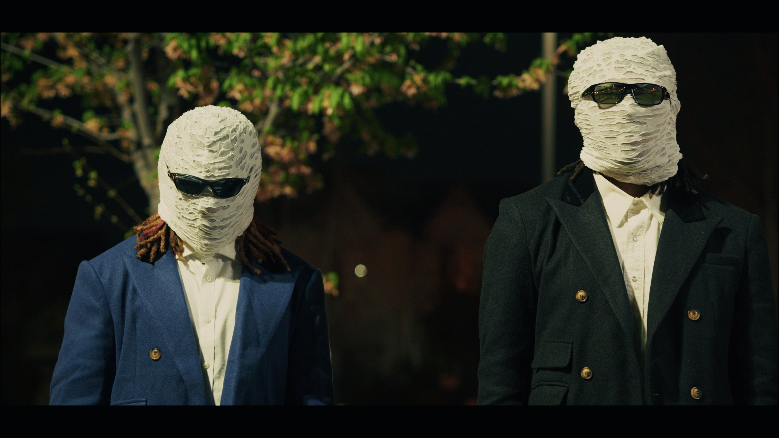 Two individuals wearing cream-colored, textured masks, sunglasses, and formal suits standing outdoors at night.