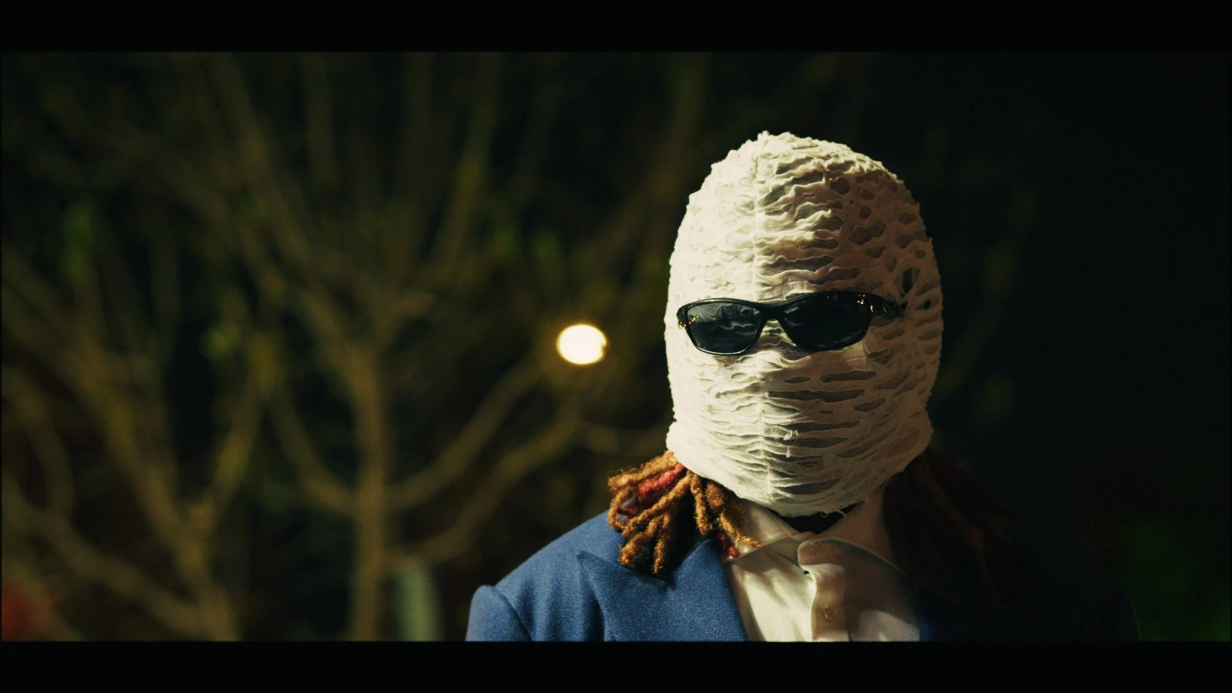 Person wearing a large white textured mask and sunglasses, dressed in a blue jacket and white shirt, standing outdoors at night.