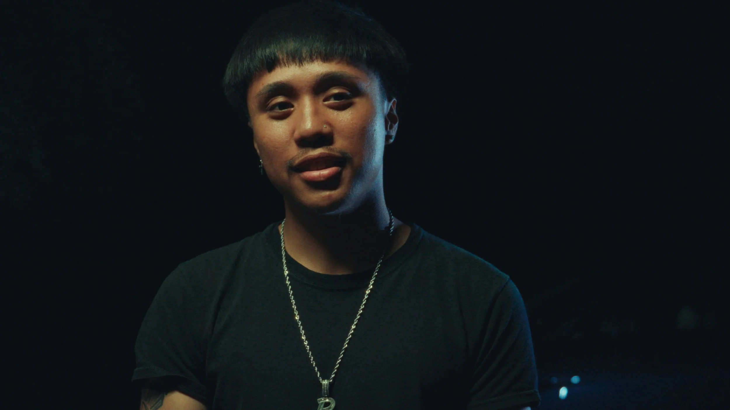 A young man with short black hair, a nose piercing, wearing a black shirt and a silver chain with a pendant, against a dark background.