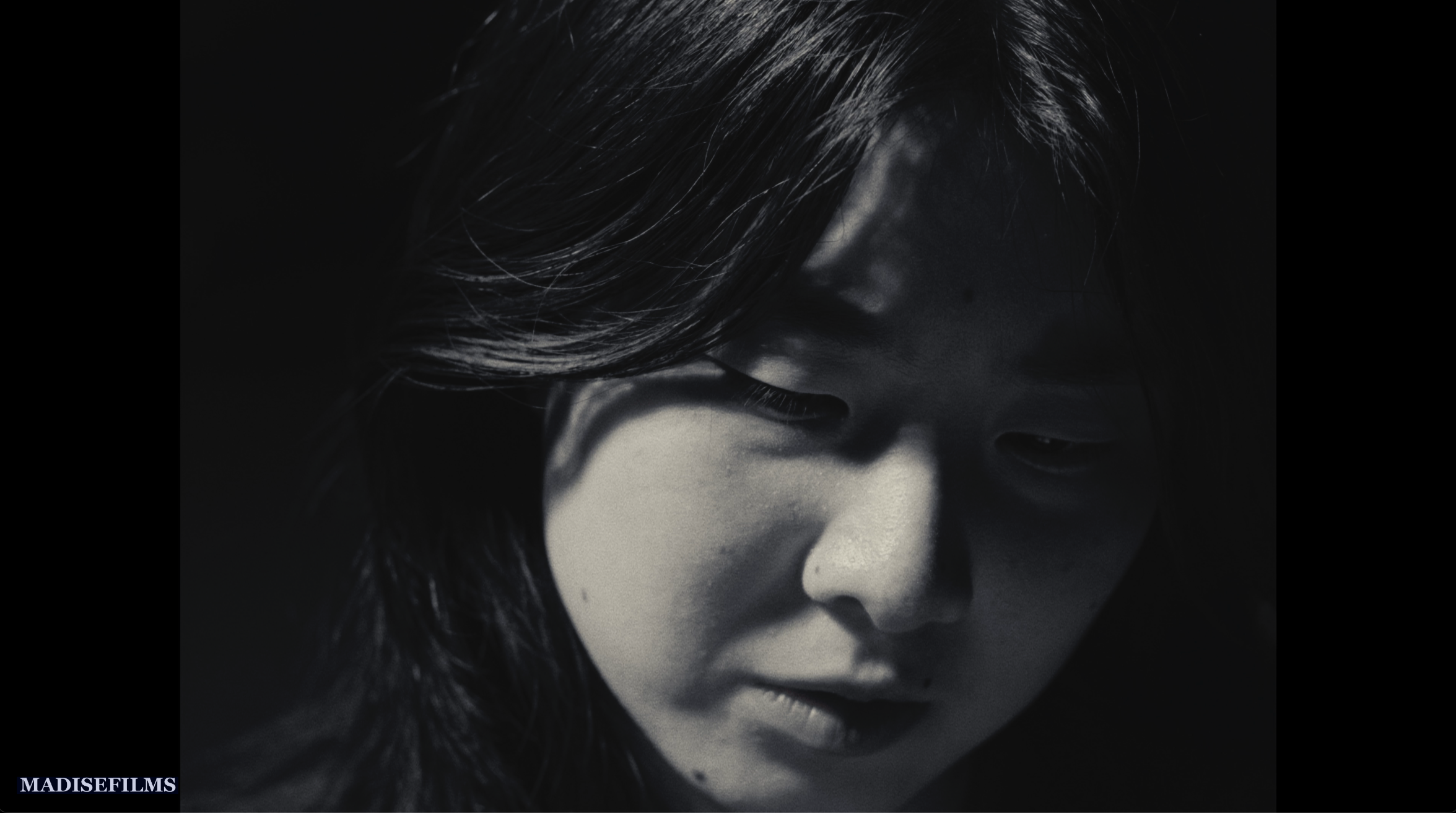 Close-up of a person's face with long dark hair, looking downward, in black and white.