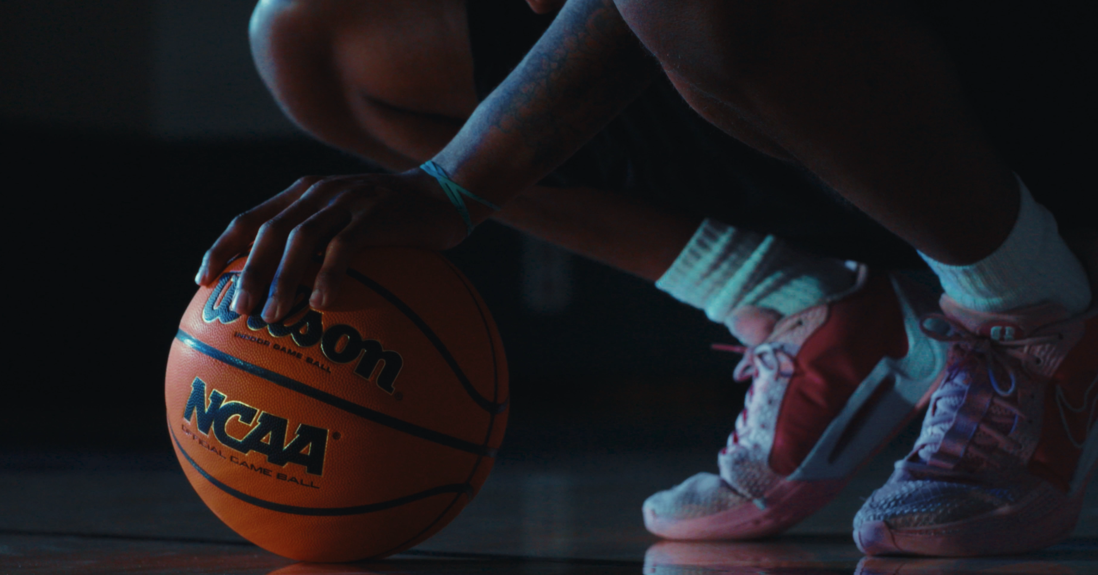 A person in athletic shorts and running shoes is placing their hand on a basketball on a dark surface.