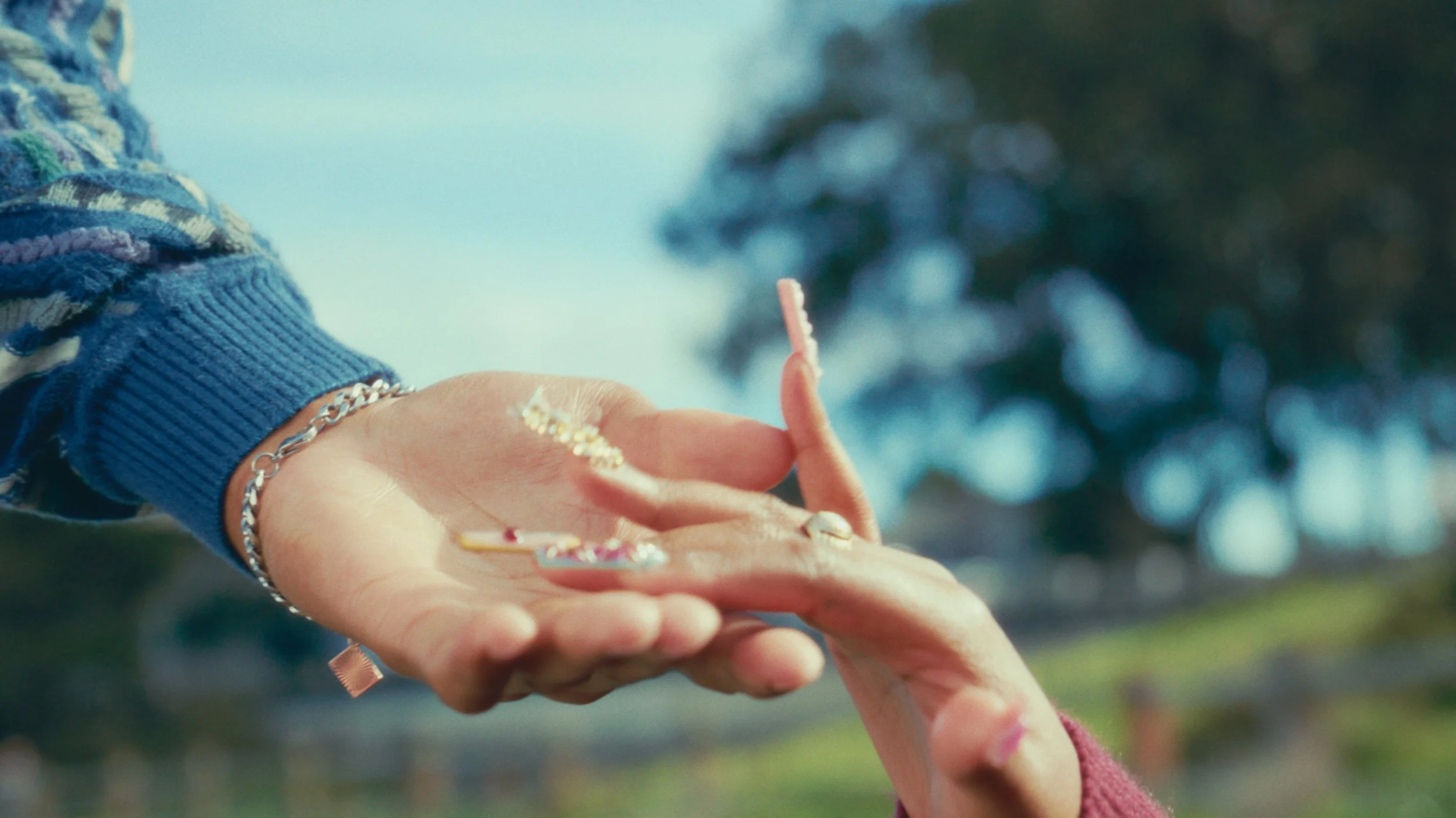 Two hands, one with jewelry, hold a small object with a stick or tool to a child's hand, in an outdoor setting with blurred trees in the background.