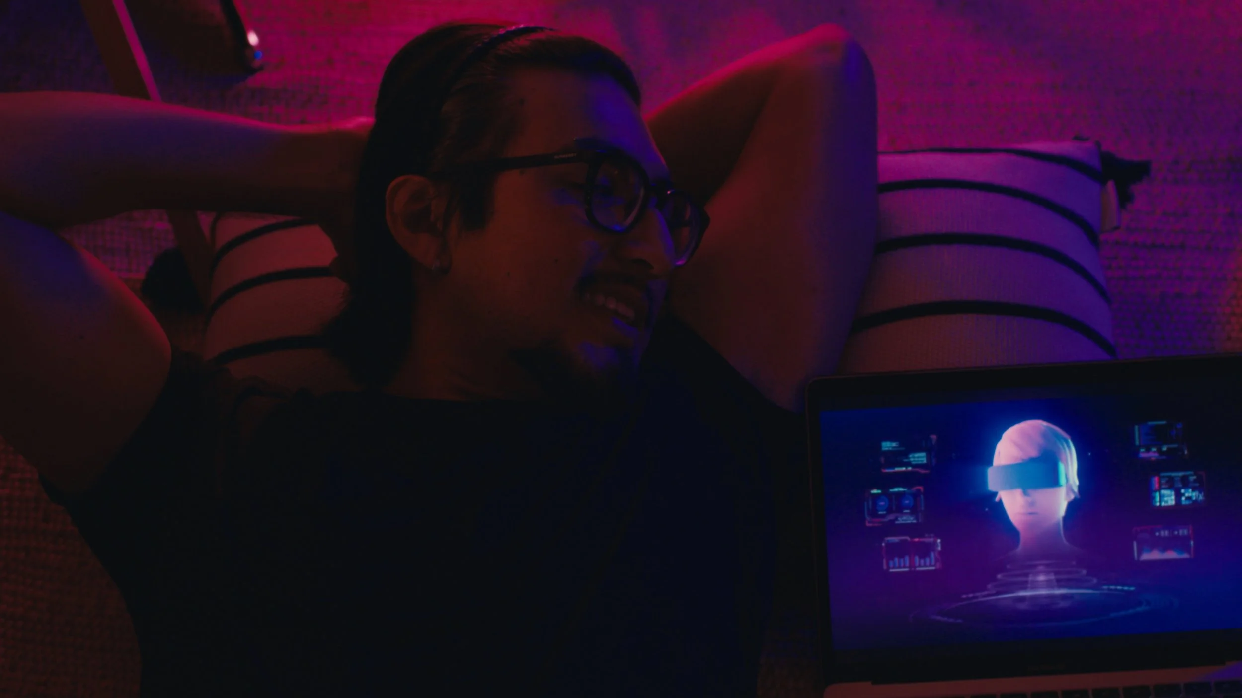 A woman wearing glasses and with dark hair is lying on a bed, smiling, while looking at a laptop screen showing a female avatar or virtual personality in a digital environment, with a mix of purple and pink lighting.