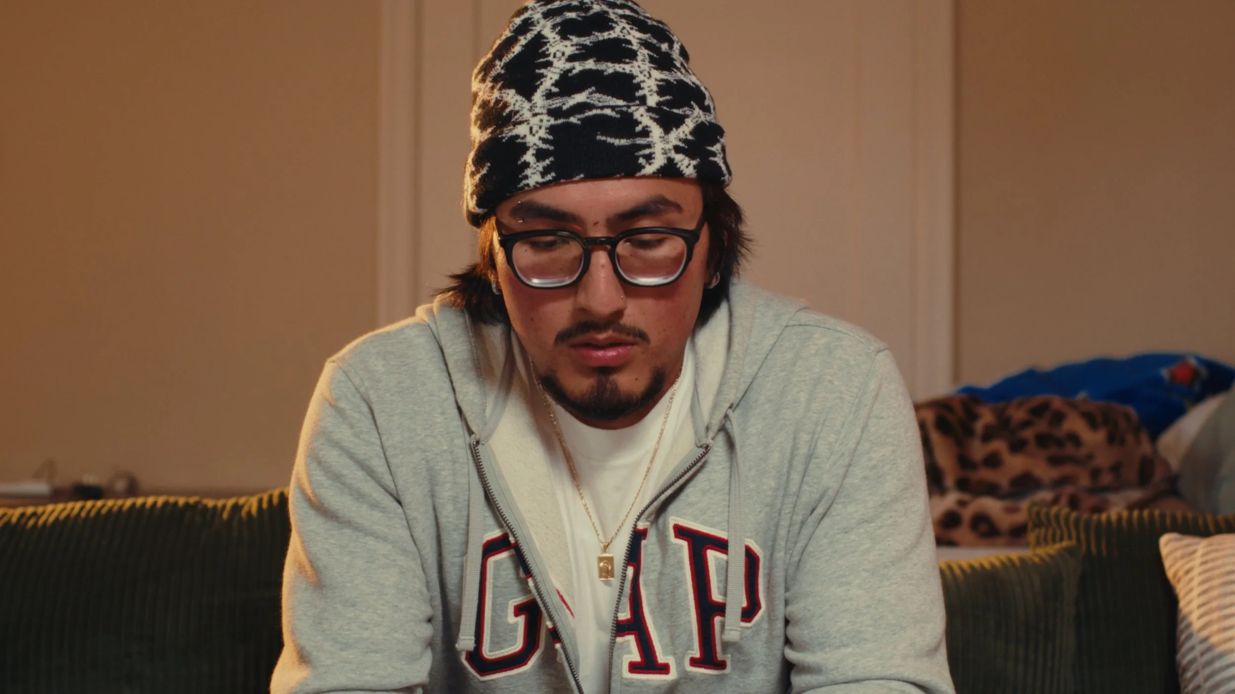 A man with glasses, a mustache and goatee, wearing a black and white patterned beanie and a gray hoodie, looking down with a serious expression in a living room with a leopard print blanket in the background.