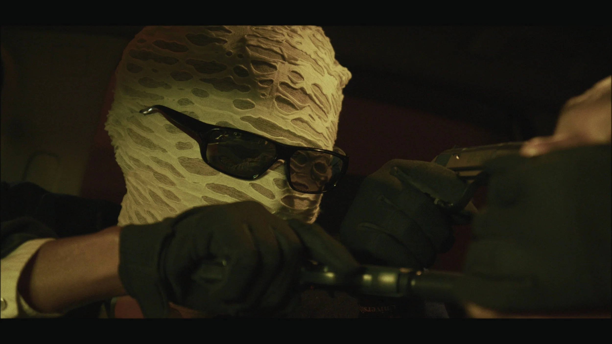 A person wearing a beige textured hoodie and black sunglasses is aiming a handgun with both hands.