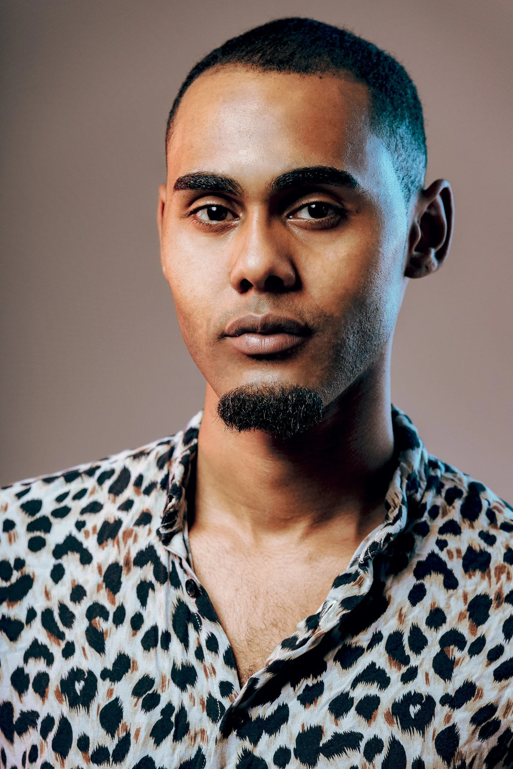 Close-up portrait of a young man with short black hair, wearing a leopard print shirt with the top buttons undone, against a plain background.