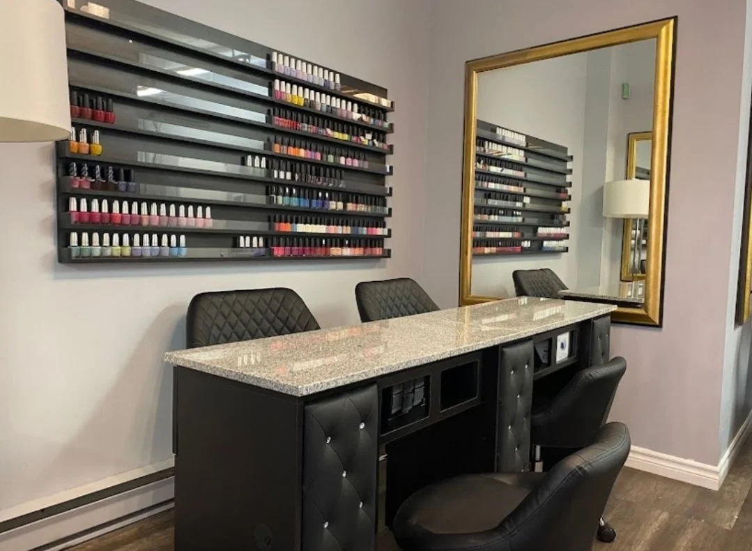 A nail salon workspace with a granite countertop, three black tufted chairs, a large gold-framed mirror on the wall, a wall-mounted nail polish display with multiple rows of colorful nail polish bottles, and a floor lamp reflected in the mirror.