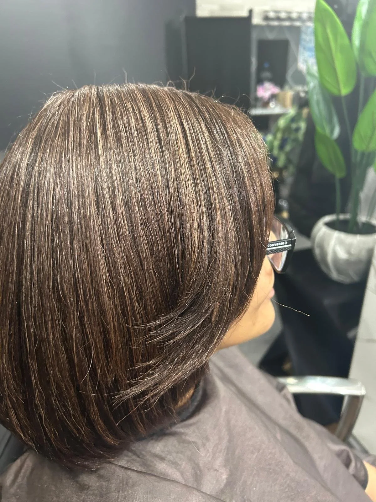 A woman with shoulder-length brown hair, wearing glasses, is sitting in a salon chair. She appears to be getting her hair styled or treated, with a large potted plant and salon equipment visible in the background.