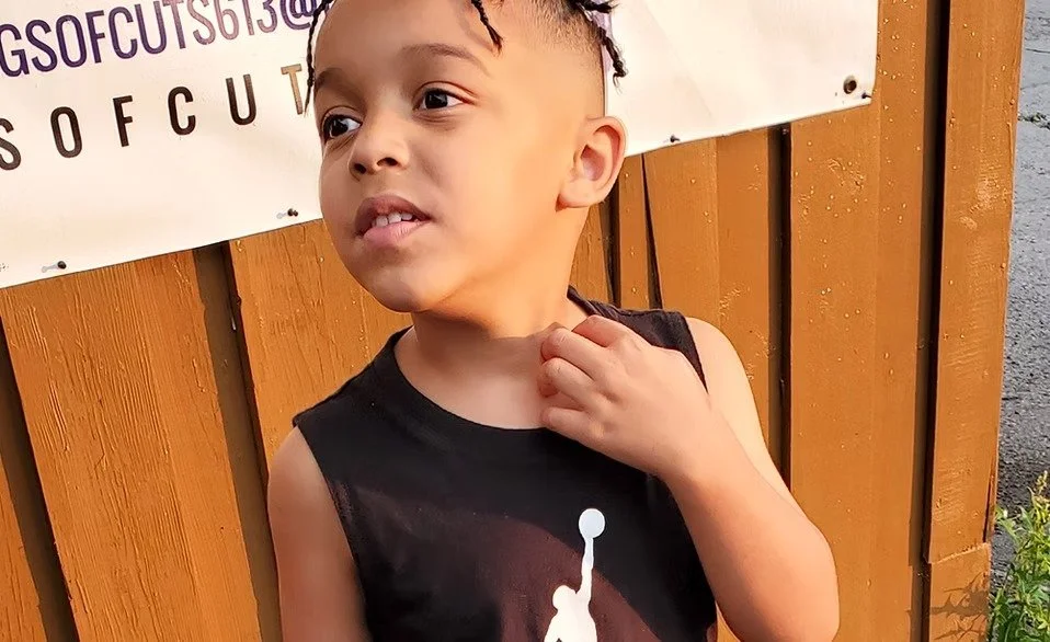 A young boy with short curly hair wearing a black sleeveless shirt, standing outdoors near a wooden wall and looking to the side.