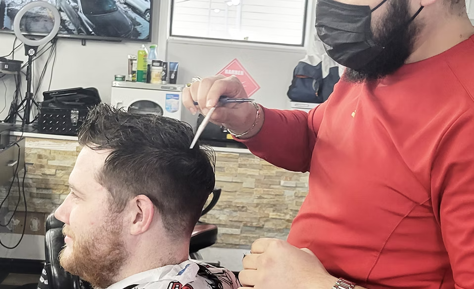 Barber in red shirt and black face mask giving a male customer a haircut in a barbershop, with hair clippers and styling tools visible.