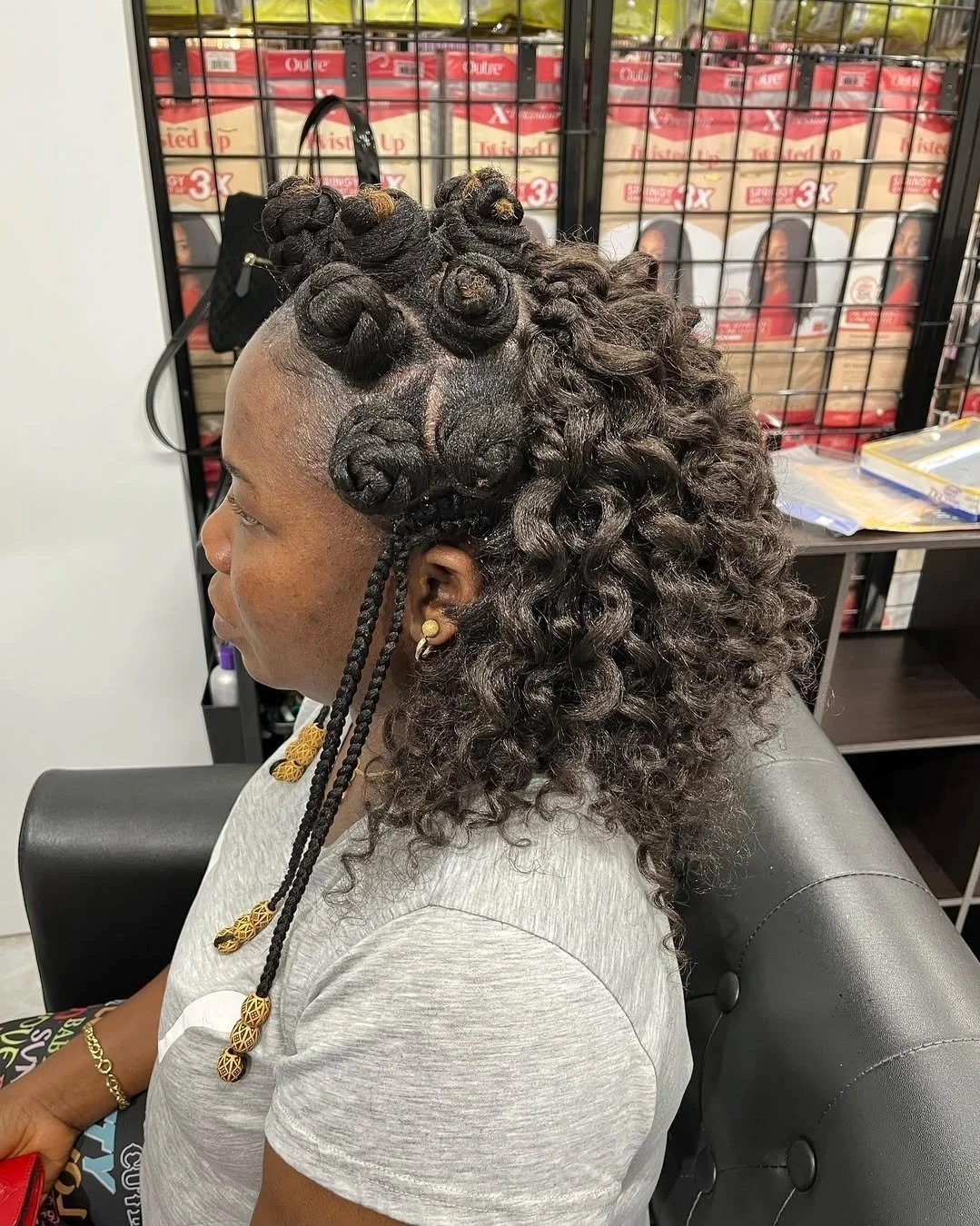 A woman with shoulder-length curly hair, some of which is styled into bantu knots with extensions that are decorated with gold beads, sitting in a salon chair. She is wearing a gray T-shirt, gold earrings, and a gold bracelet. The background shows sh