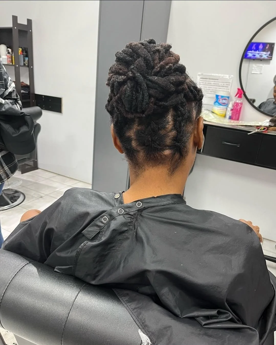 Back view of a woman with styled curly dreadlocks, sitting in a salon chair, wearing a black cape.