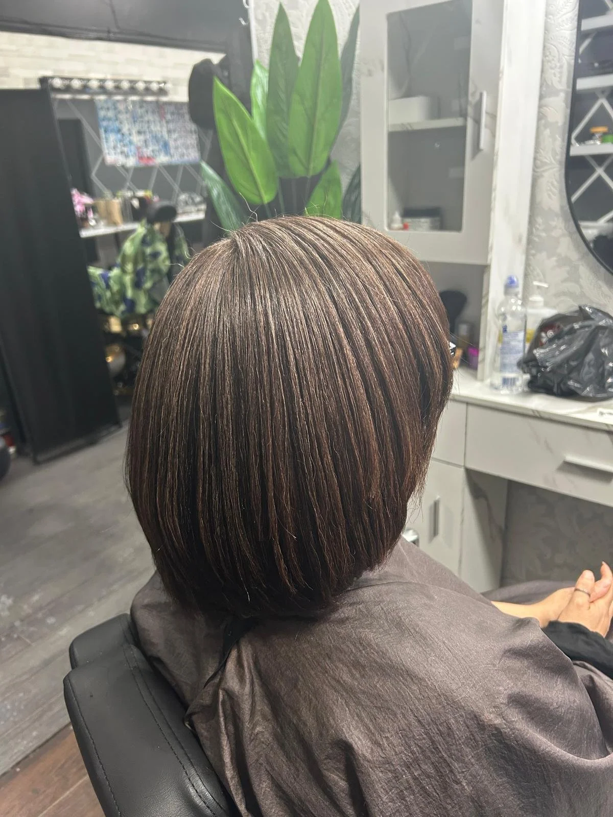 A woman with shoulder-length, shiny brown hair with lighter highlights, sitting in a salon chair in a hair salon.