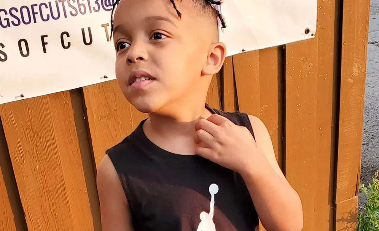 A young boy with braided hair standing outdoors in front of a wooden fence and a white banner, wearing a sleeveless black shirt.