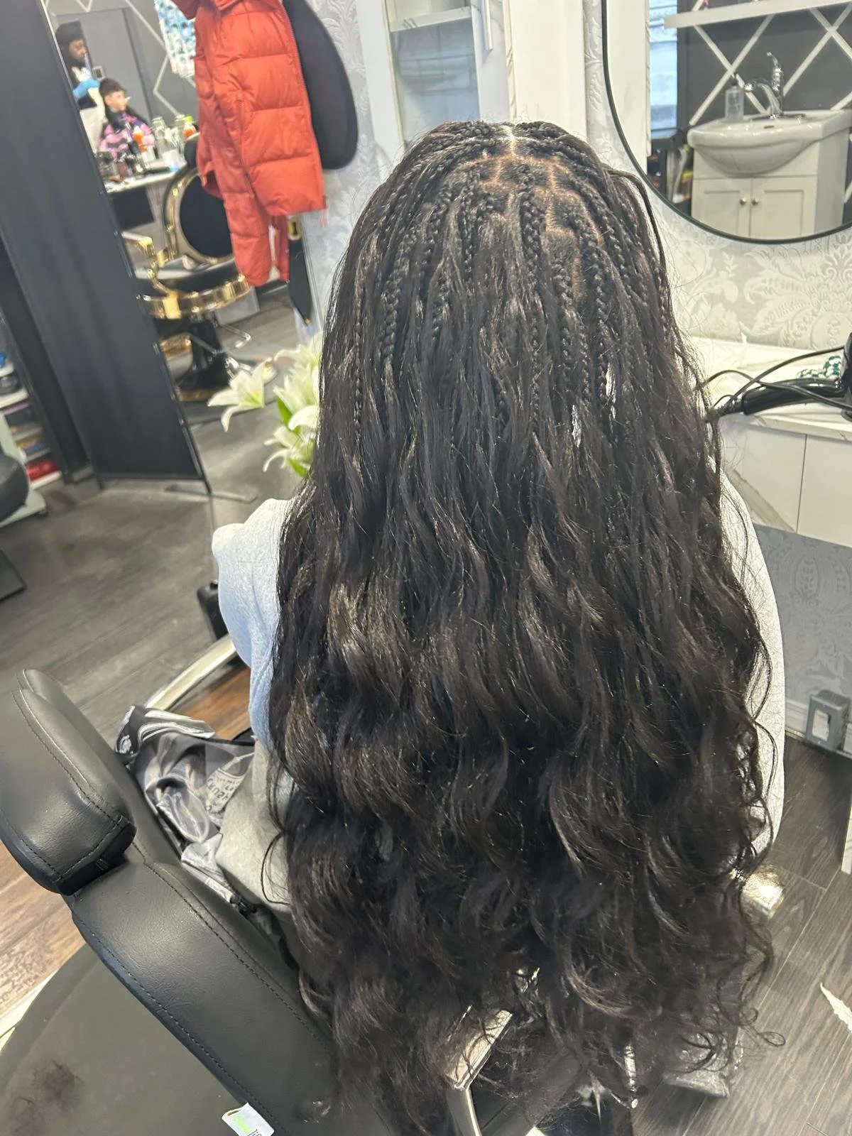 A person with long, curly black hair sitting in a salon chair in front of a mirror, with salon equipment and other people visible in the background.