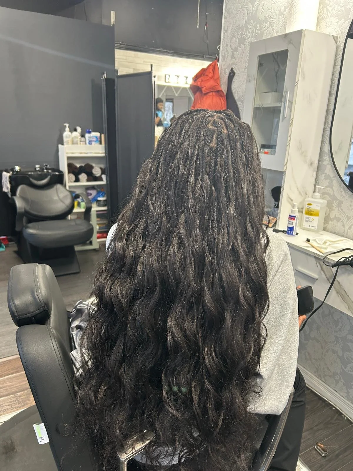 Back of a woman with long, curly, dark hair sitting in a salon chair at a hair salon, with hair tools and products on the counter in front of her.