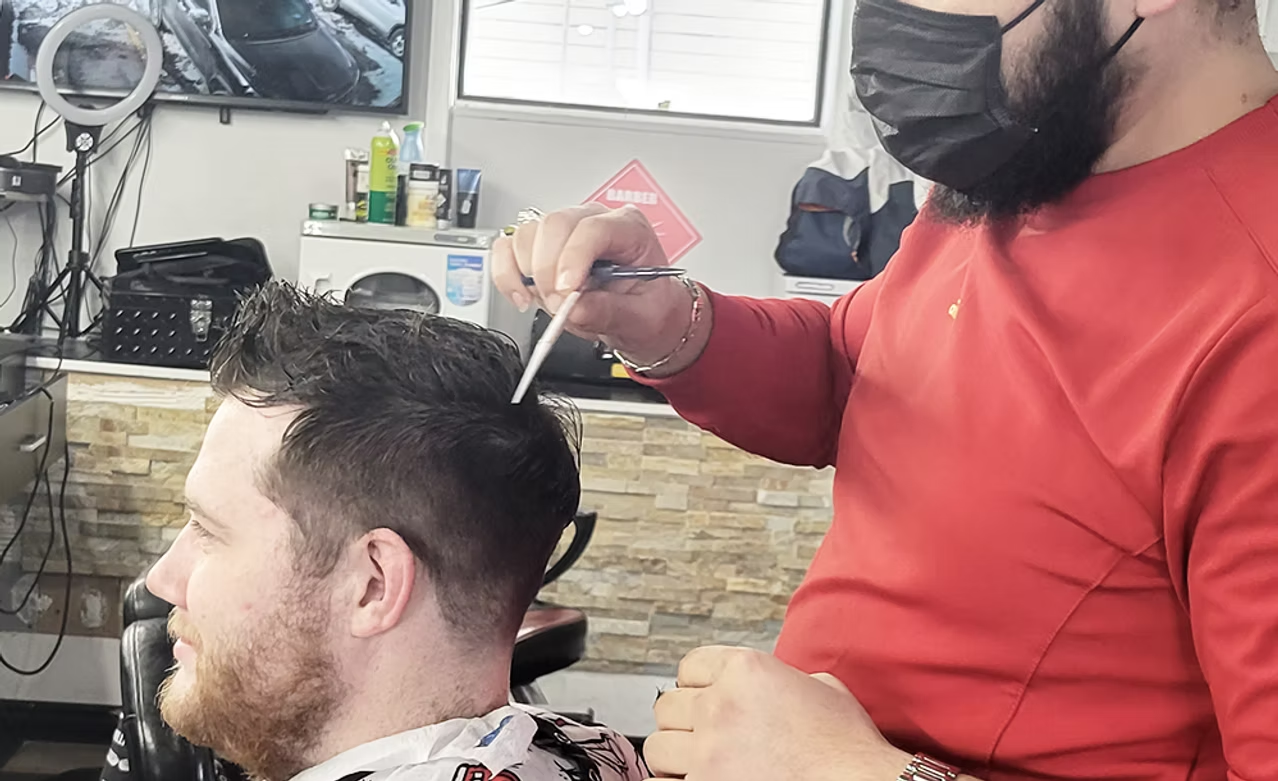 Barber cutting young man's hair in a barbershop, with barber wearing a red shirt and a black face mask.