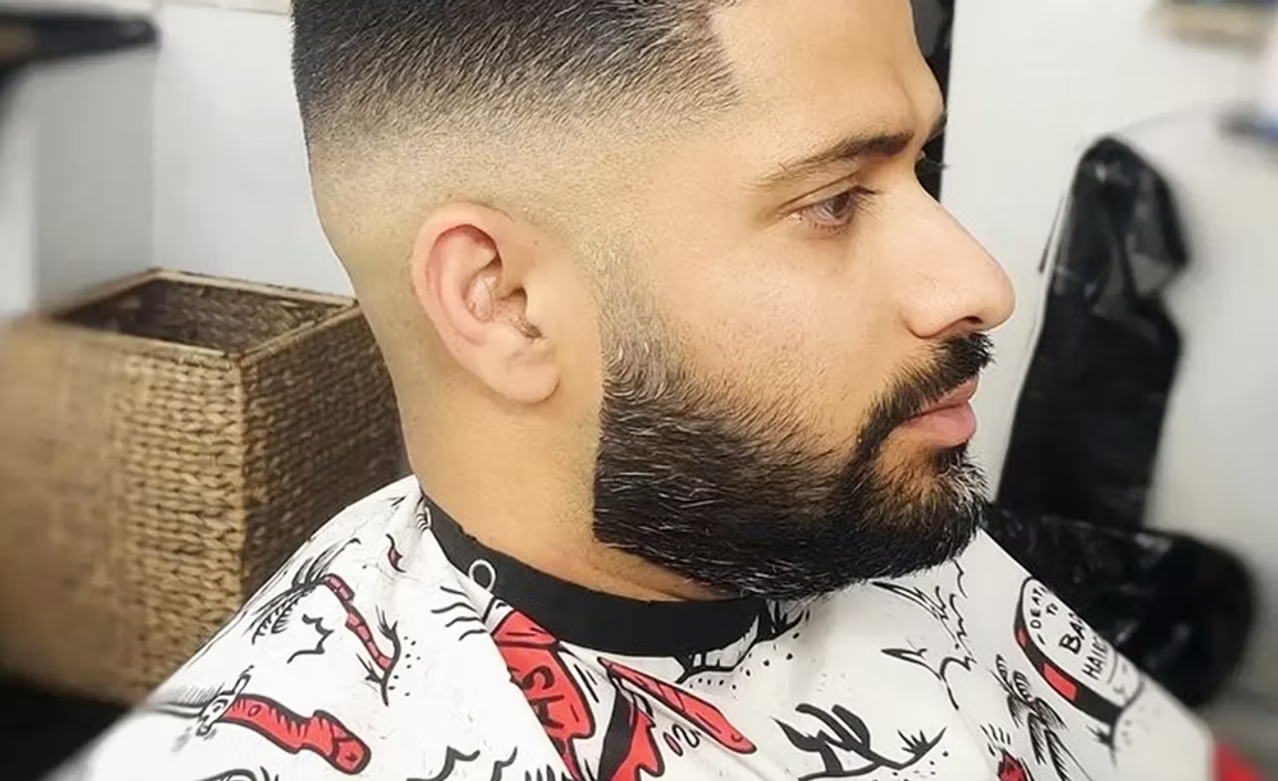 Side view of a man with a fresh fade haircut and a thick, well-groomed beard, sitting in a barbershop.