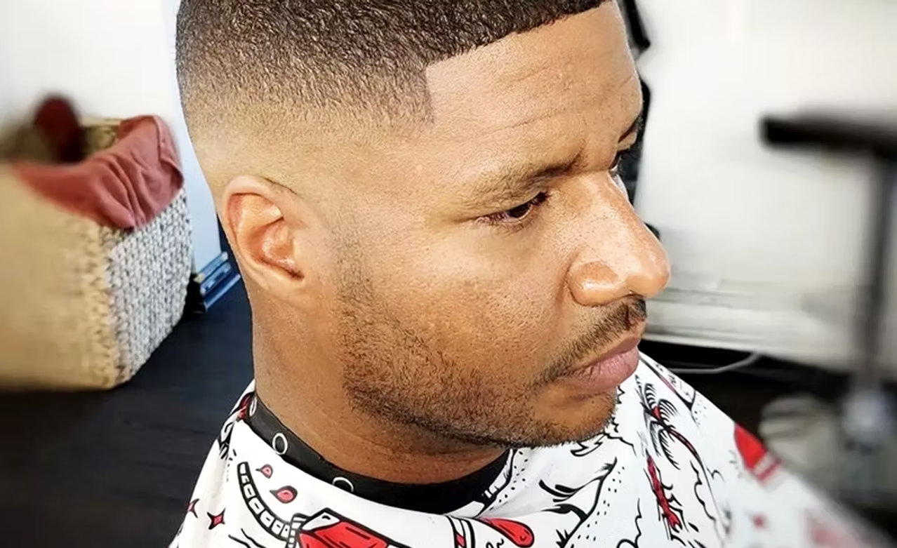 Close-up of a man with a fresh buzz cut hairstyle, sitting in a barber's chair, wearing a barber cape with cartoon graphics, in a salon or barbershop setting.