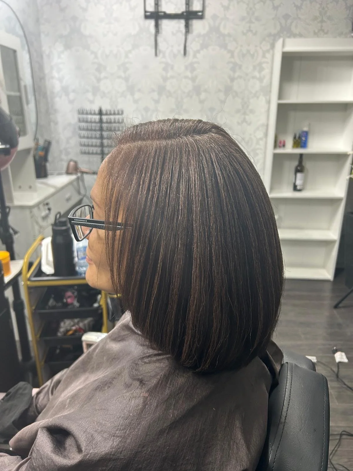 Side view of a woman with shoulder-length straight hair, brown with blonde highlights, sitting in a salon chair, wearing glasses. The background shows salon equipment and shelves.