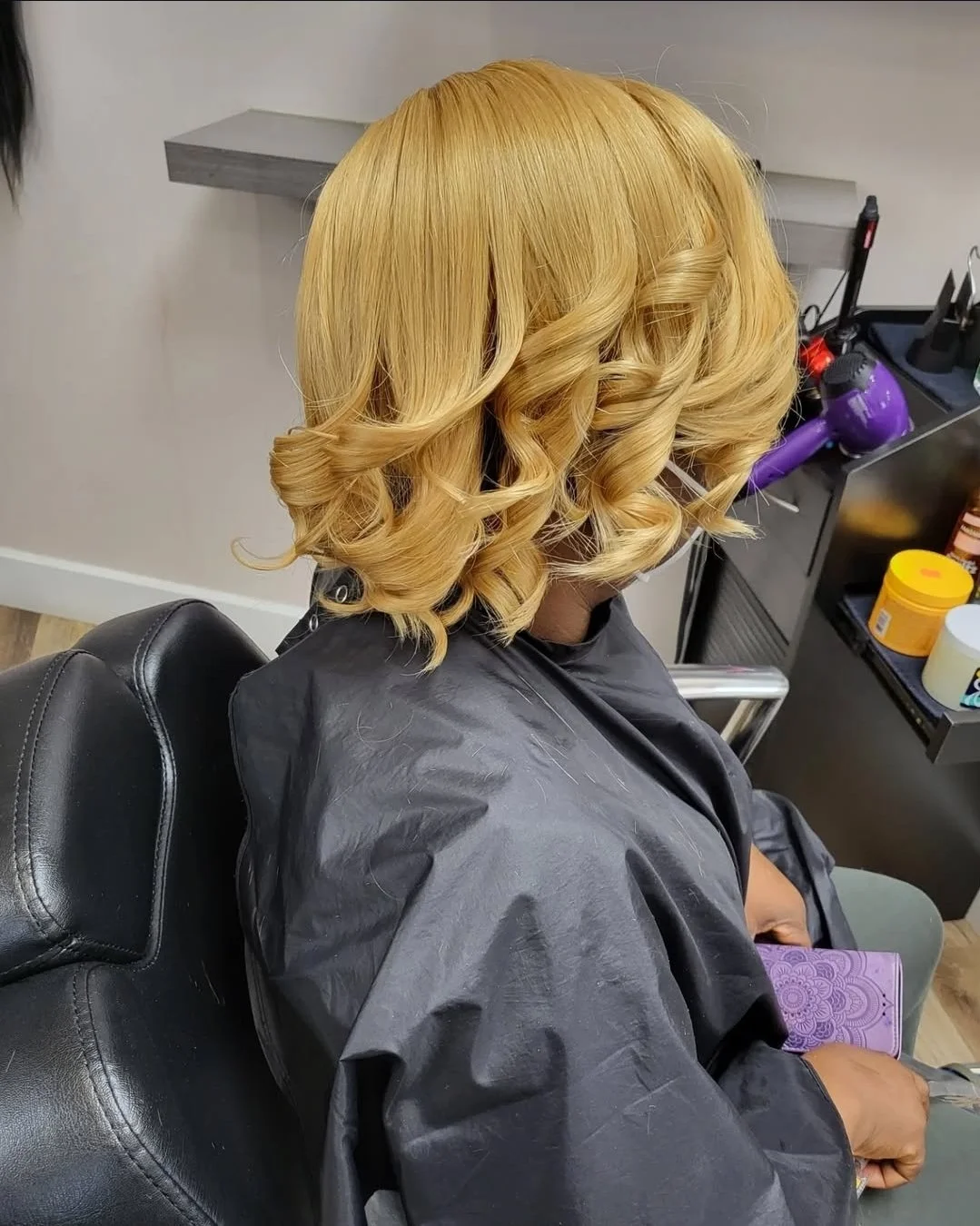 Person with blonde, curled hair sitting in a salon chair, wearing a black cape, holding a purple notebook.