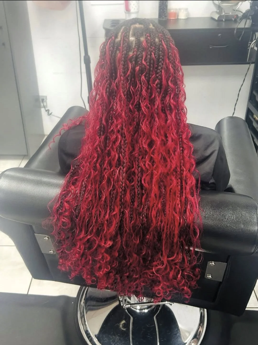Person with long, curly hair that is dark at the roots and transitions into bright red, sitting in a black salon chair in a hair salon.
