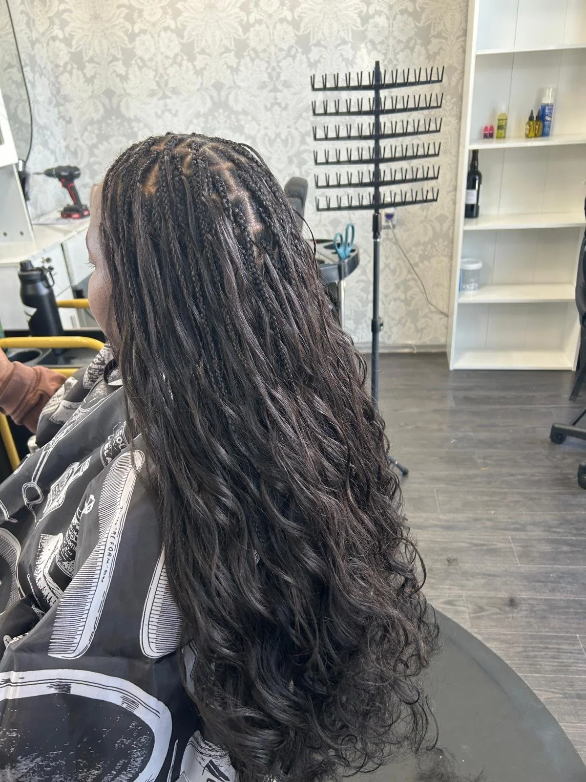 Woman sitting in a salon chair, getting her hair styled with braids and loose curls, in a salon with shelves and hair products in the background.