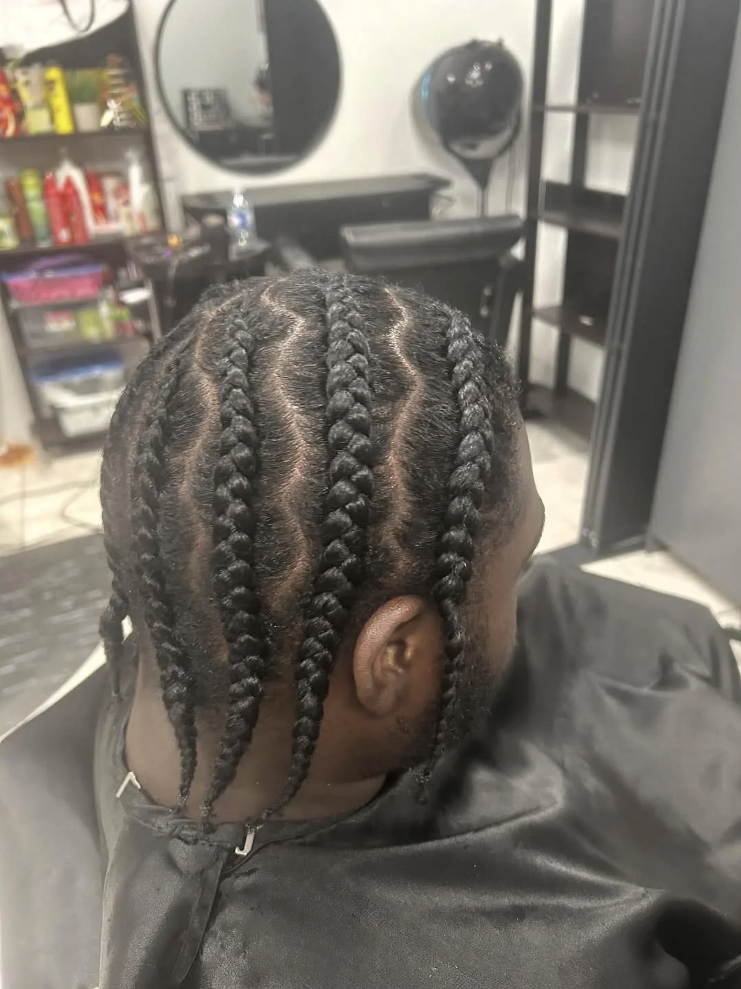 A person with freshly styled hair in neat, thick twists, sitting in a salon chair with a black cape, behind a styling station with shelves and a mirror.