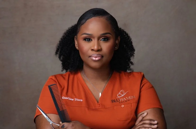 A confident woman with natural curly hair, wearing an orange uniform with embroidered text, holding a comb and scissors, posing against a neutral background.