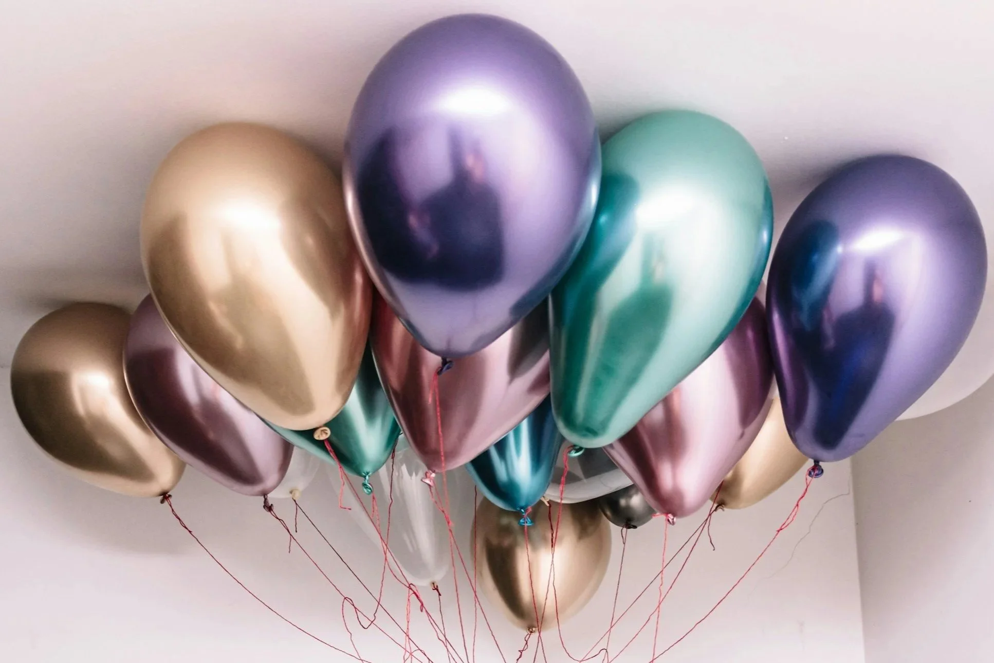 A bunch of metallic balloons in gold, purple, and teal colors are grouped together and floating against a white ceiling.