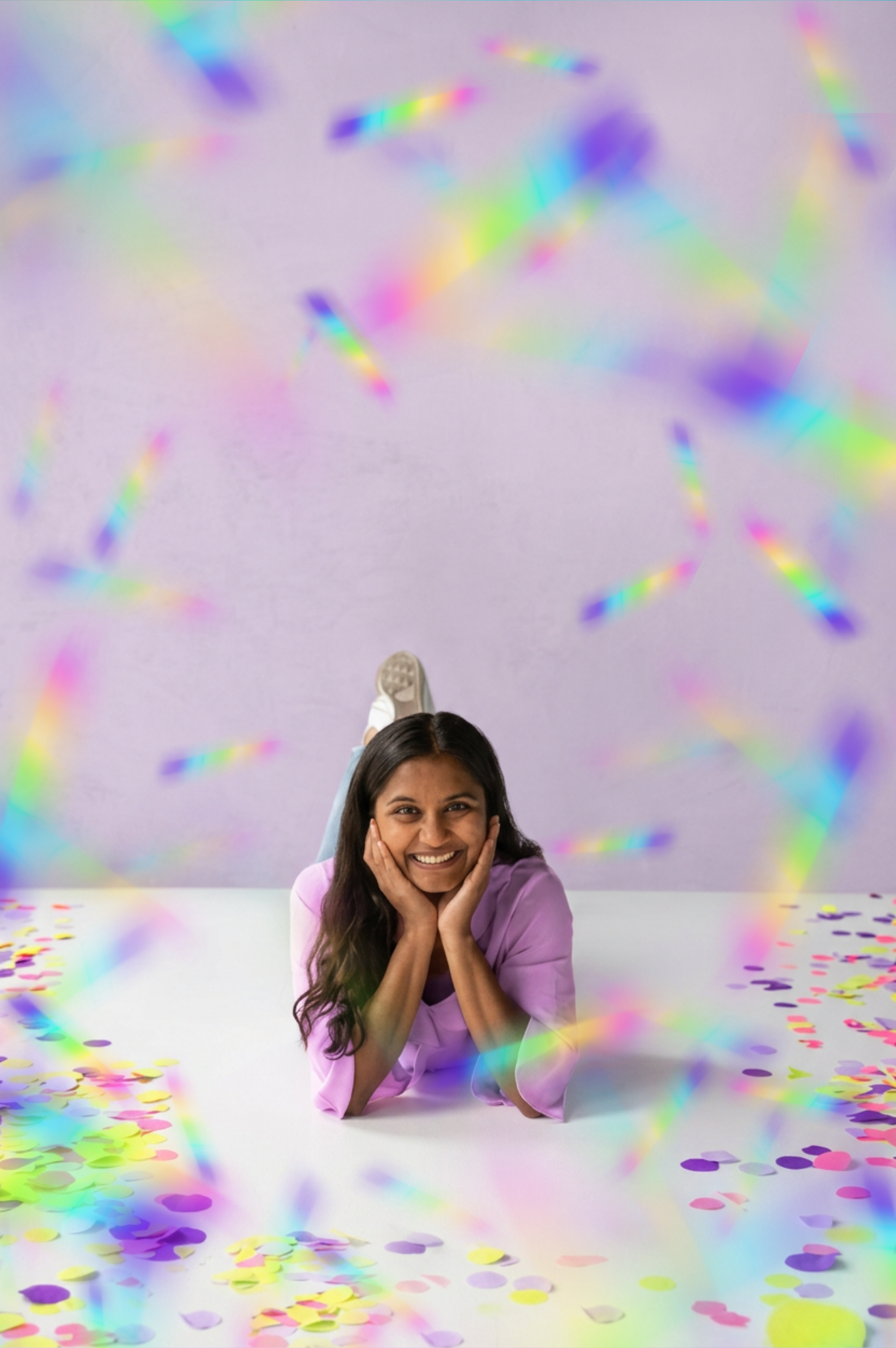 Harathi Srivastava, owner of Aura Event Designs posed with long dark hair smiling and resting her chin on her hands, sitting at a white table surrounded by colorful confetti and rainbow-colored light streaks.