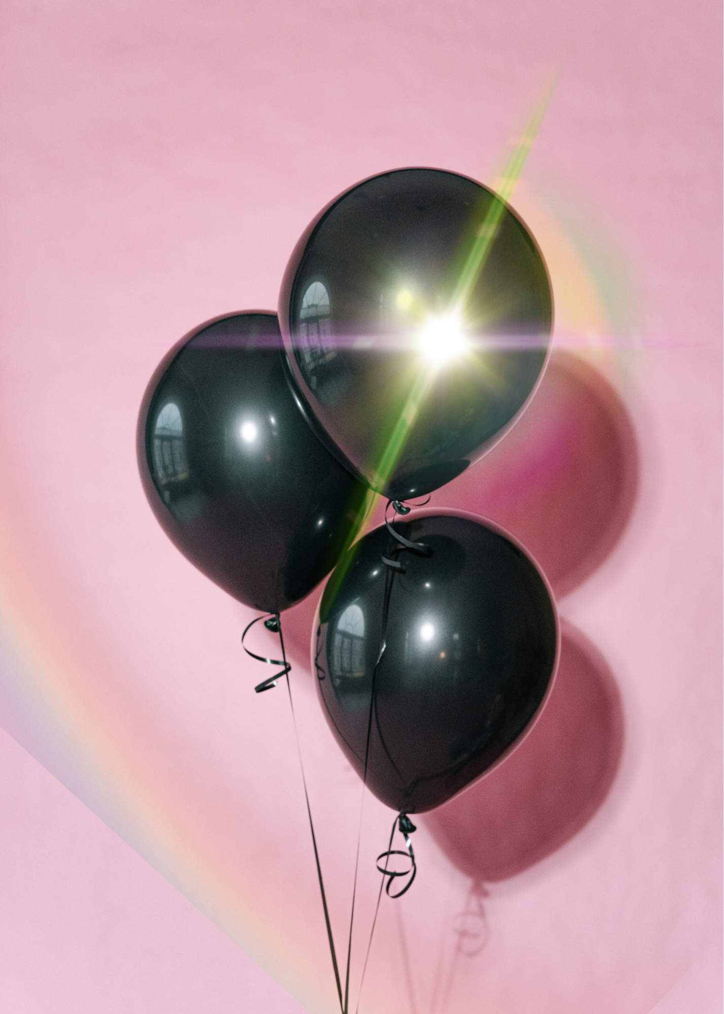 A cluster of black balloons tied with black curling ribbons, arranged against a pink background with a lens flare from the sunlight.