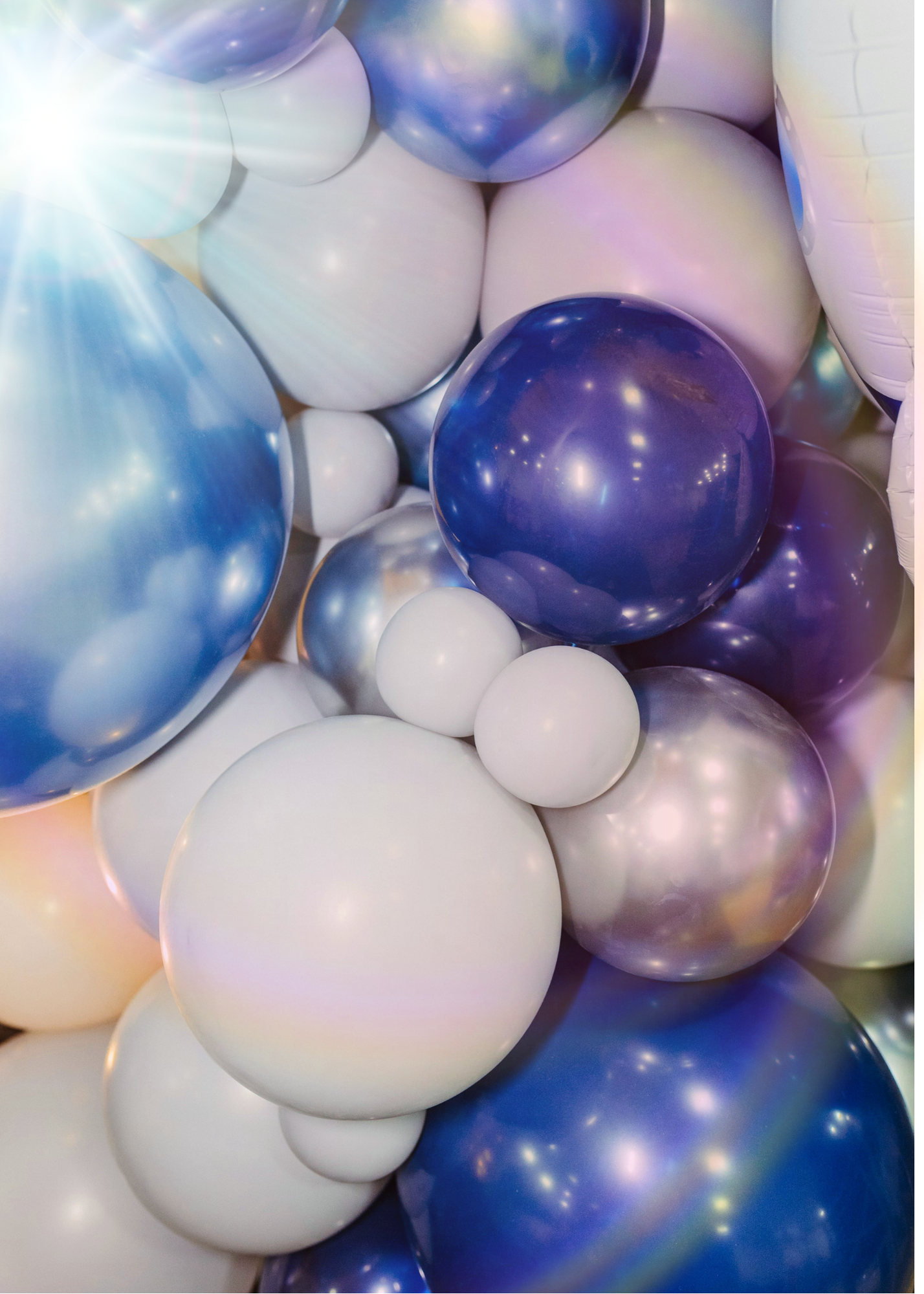A close-up view of a cluster of balloons in white, blue, purple, and iridescent colors, with sunlight reflecting off some of the balloons and creating a lens flare effect in the top left corner.
