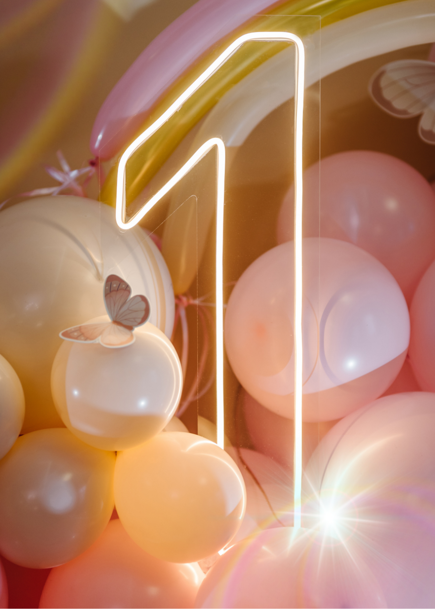 Pink, yellow, and white balloons arranged in a cluster with a neon light letter 'D' and butterfly stickers on some balloons.
