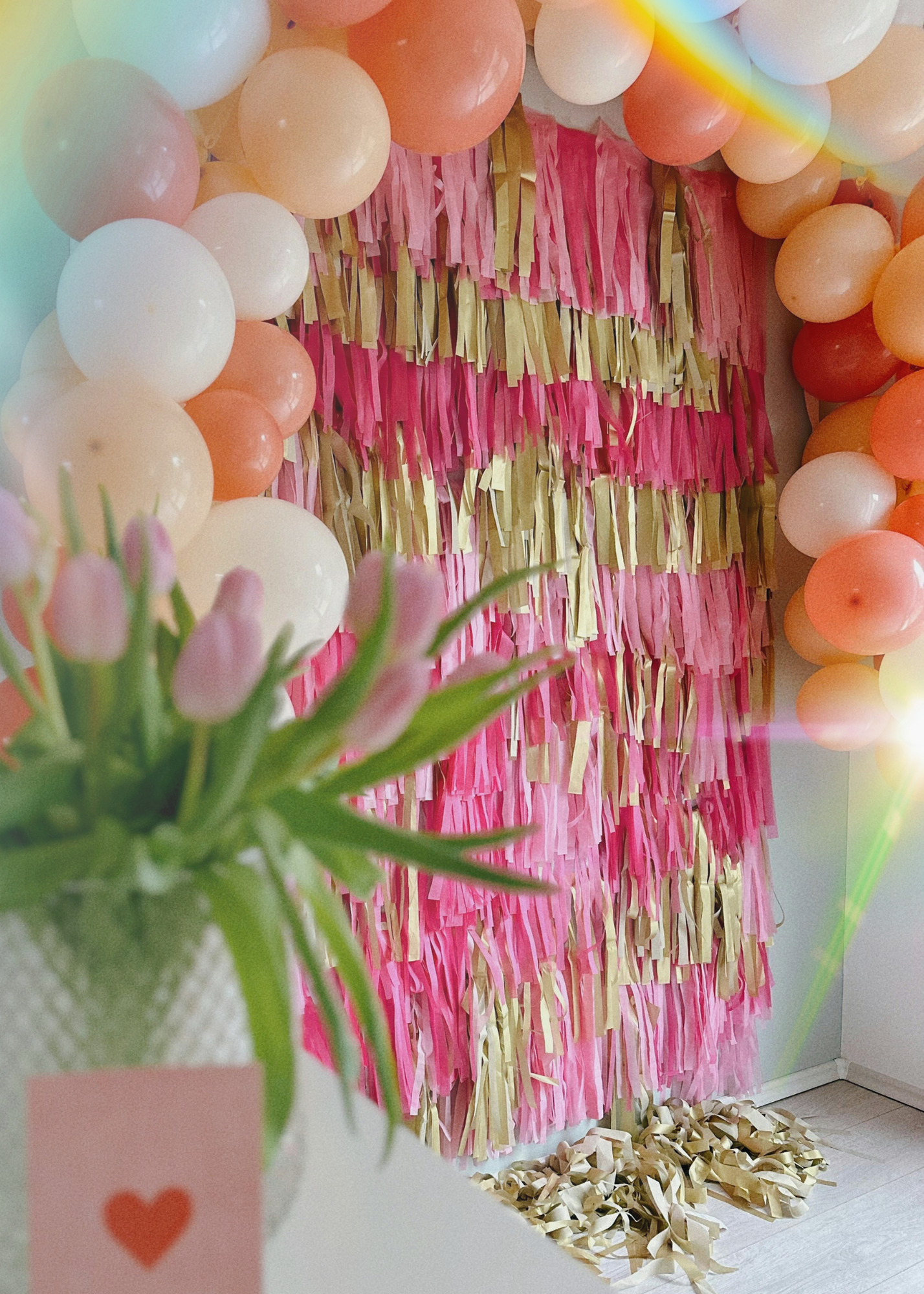 Decorative pink, cream, and orange balloons around a pink and gold fringed backdrop, with scattered paper strips on the floor, part of a floral arrangement with pink tulips in the foreground.