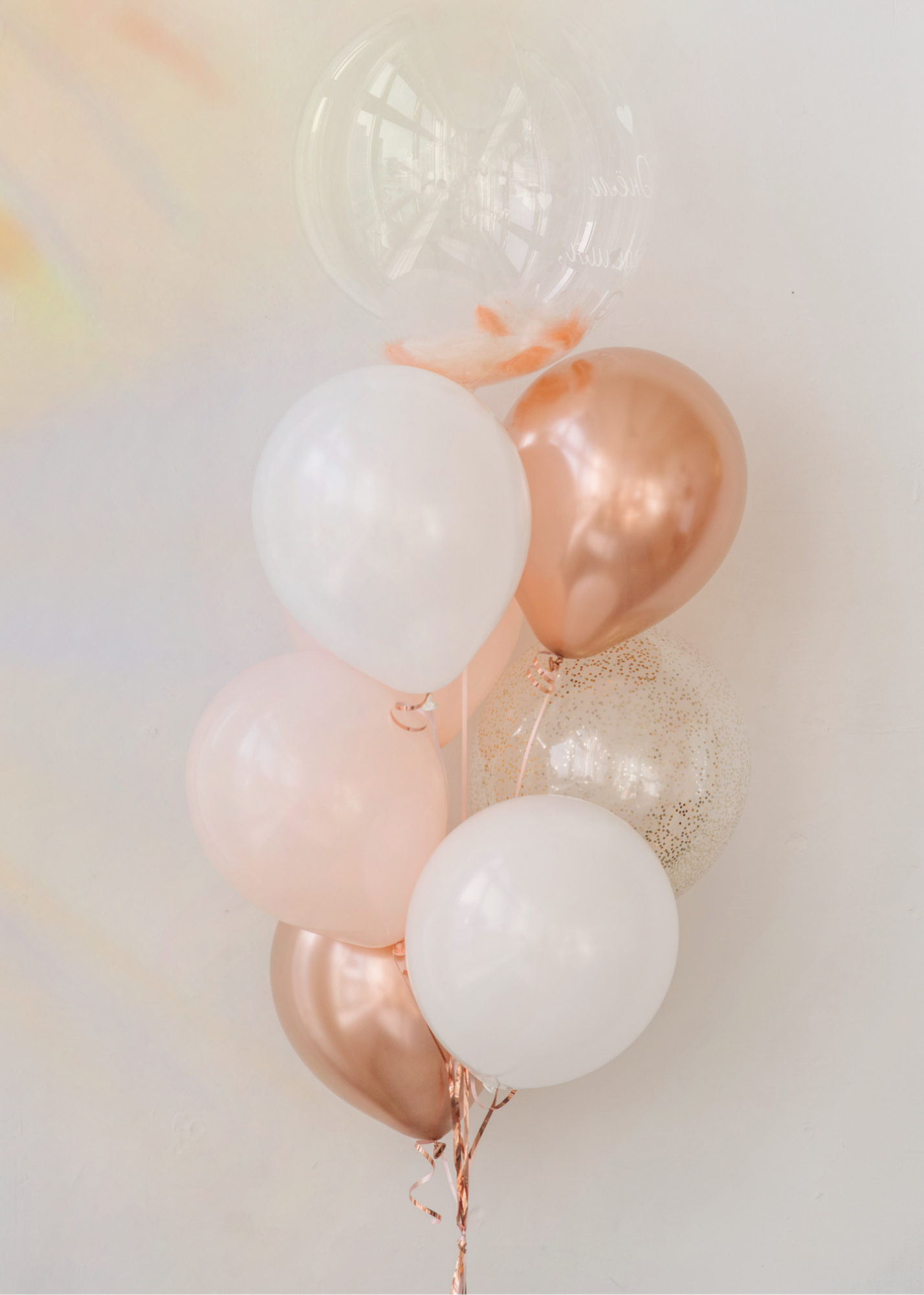 A cluster of pink, white, and transparent balloons with gold confetti and ribbon decorations, set against a light background.