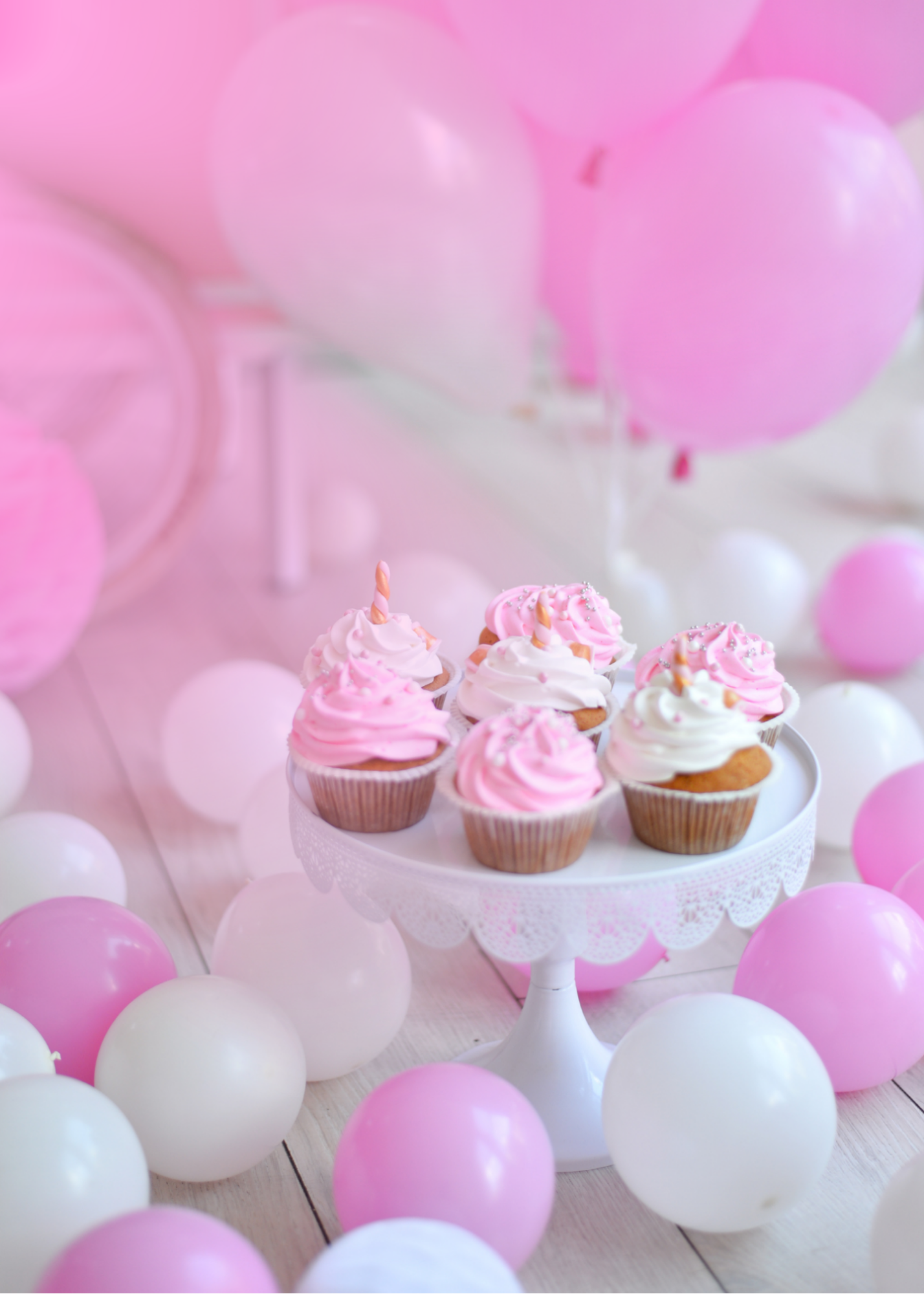 Pink and white balloons surrounding a white cake stand with pink and white cupcakes topped with pink frosting and small decorative sprinkles.