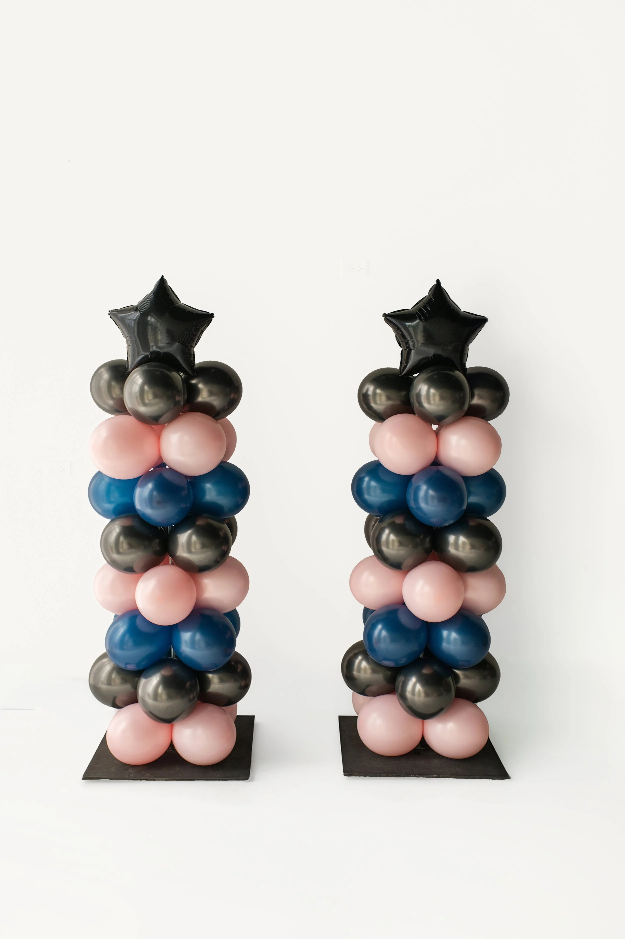 Two balloon columns topped with black star-shaped balloons, arranged with pink, blue, gray, and black balloons on black bases, against a white background.