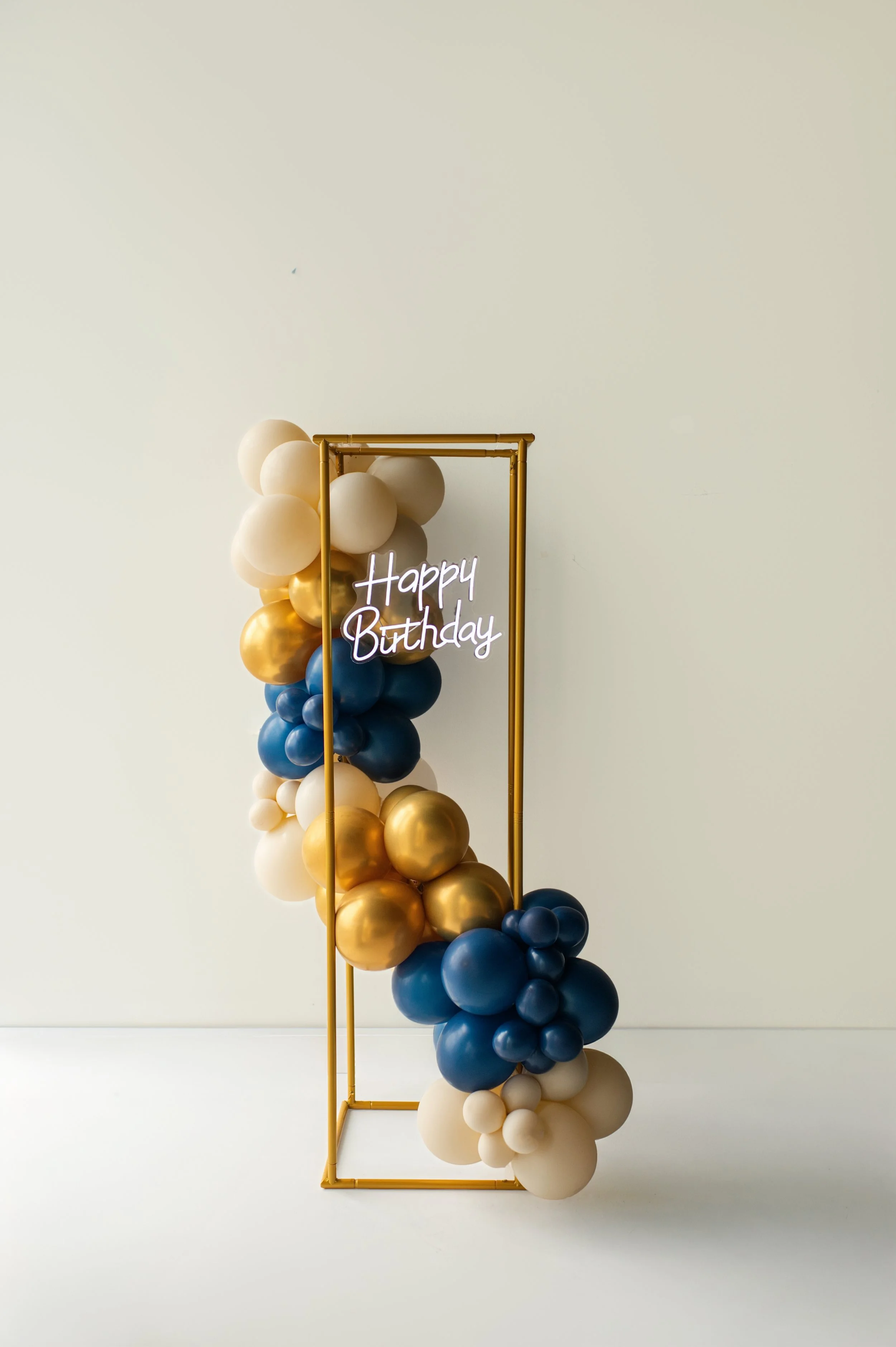 Balloon display with white, gold, dark blue, and cream balloons, gold frame, and neon sign reading 'Happy Birthday' on a plain white wall.